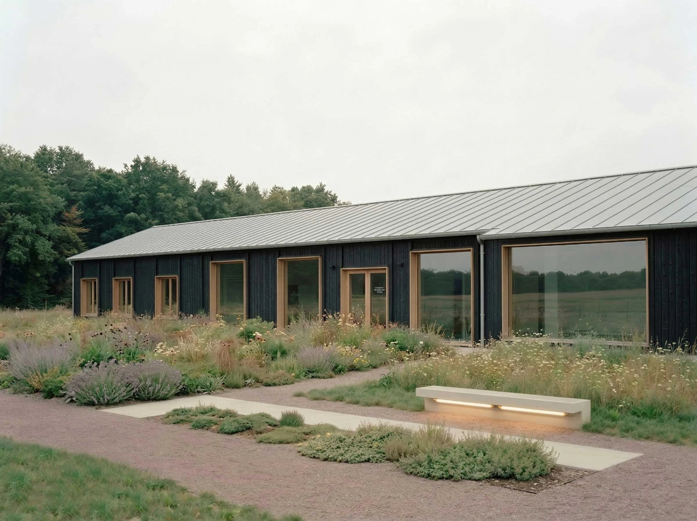 Une bâtiment moderne en bois sombre avec de grandes fenêtres, entouré de plantes et de sentiers dans un environnement naturel avec des arbres en arrière-plan.