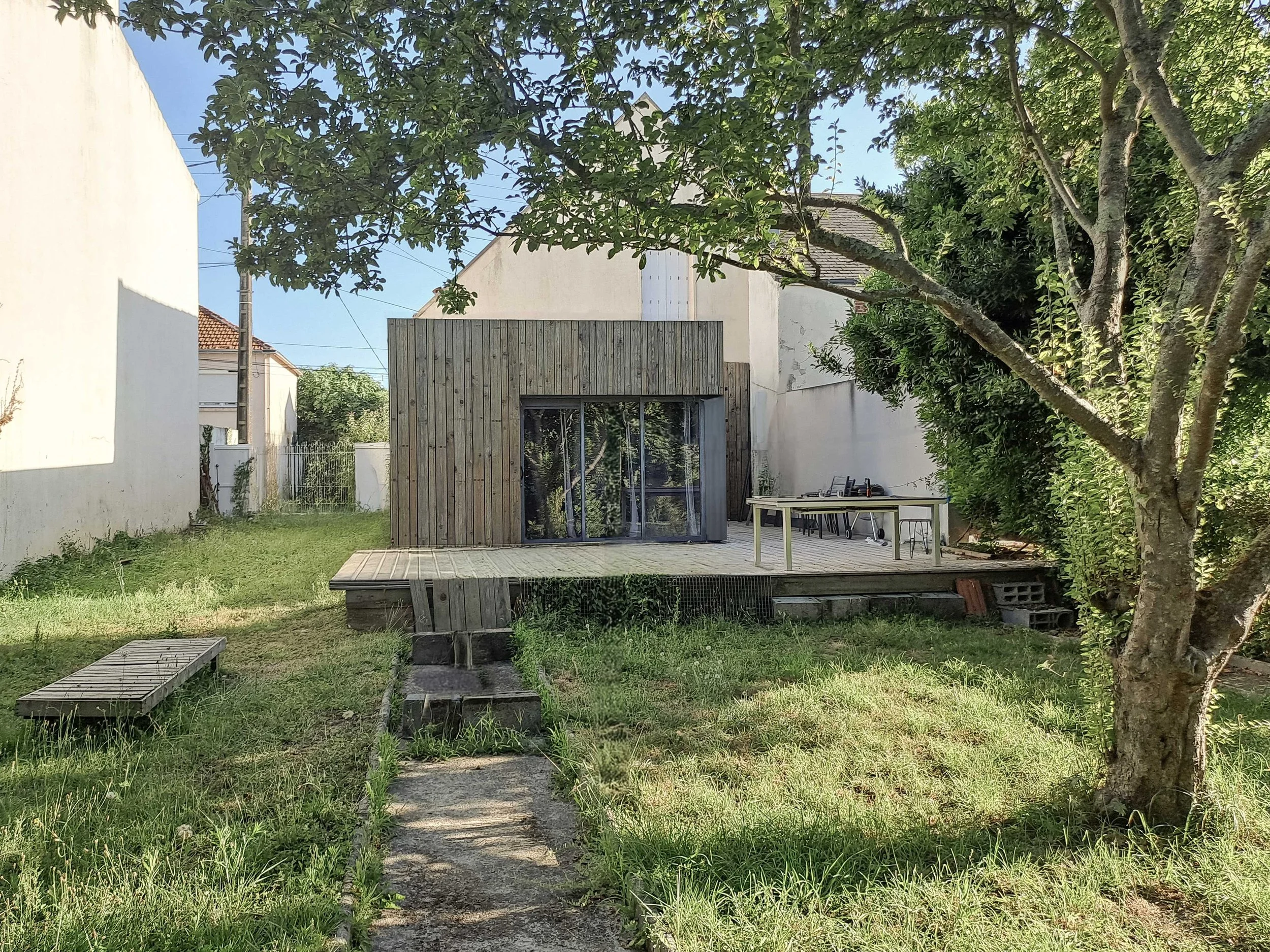 Jardin avec une pelouse verte, un arbre à droite, une structure en bois avec des fenêtres, une table et des chaises à l'arrière, un chemin en pierre au centre.