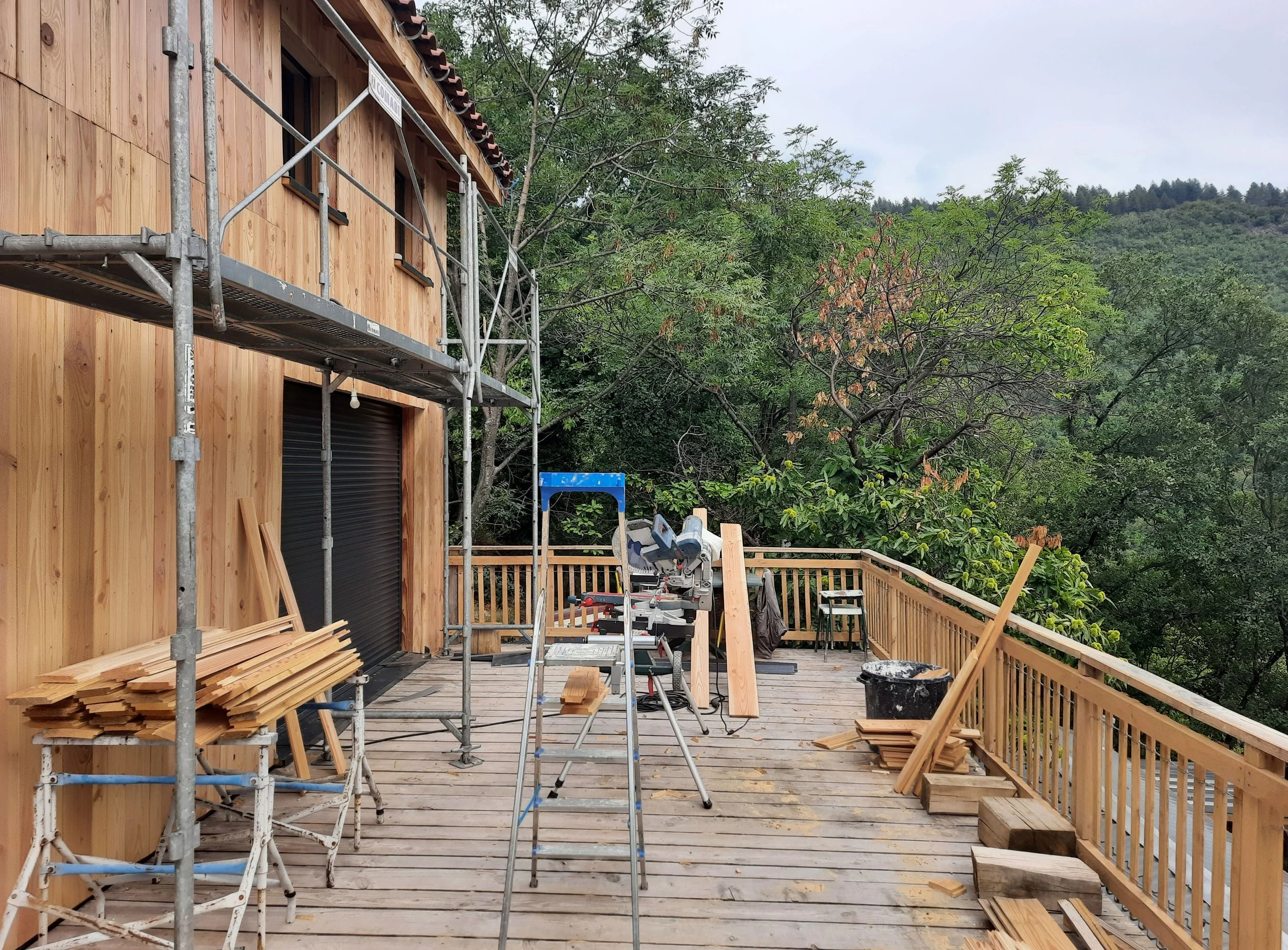 Une terrasse en bois en construction avec des outils et du bois empilé, entourée d'arbres et de montagnes en arrière-plan.