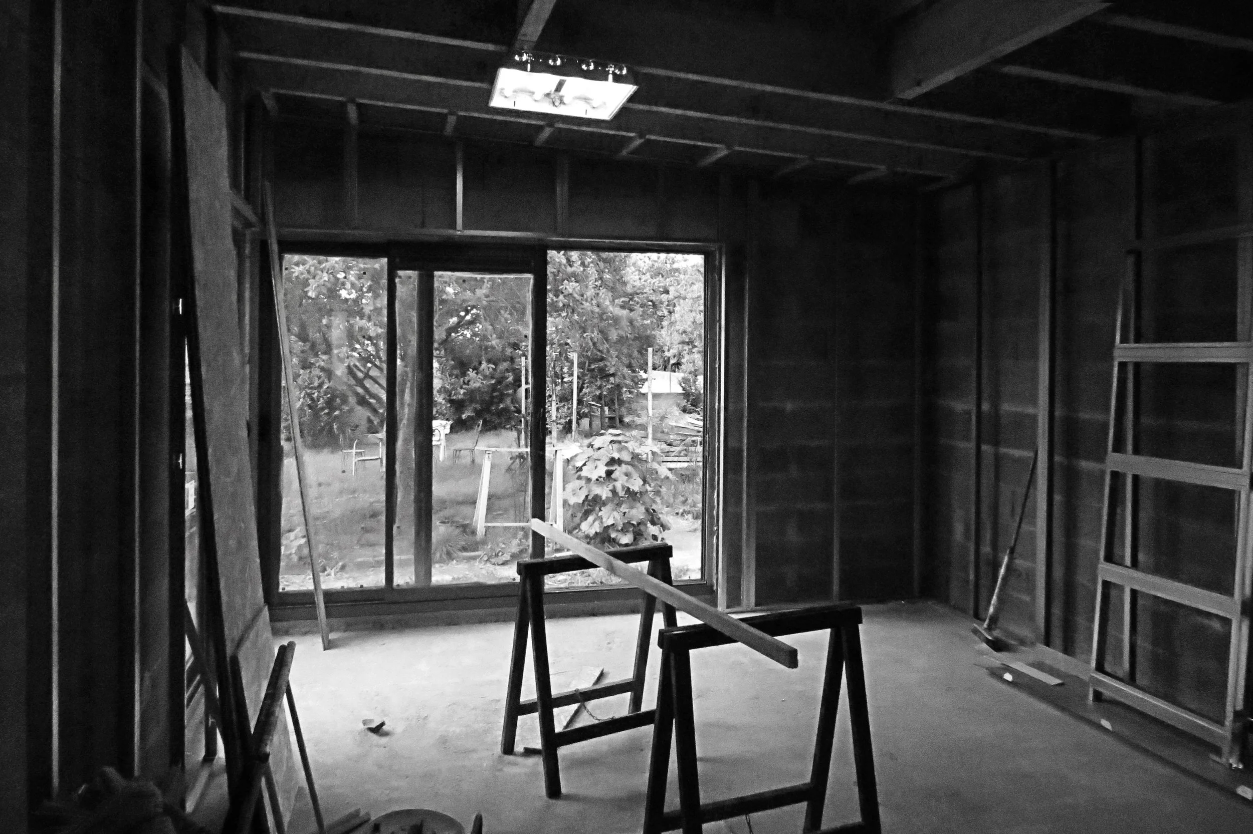 À l'intérieur d'une maison en construction, vue du salon avec des chaises en bois, une grande fenêtre donnant sur un jardin avec des arbres, mur en bois non fini, outils et matériaux de construction présents.