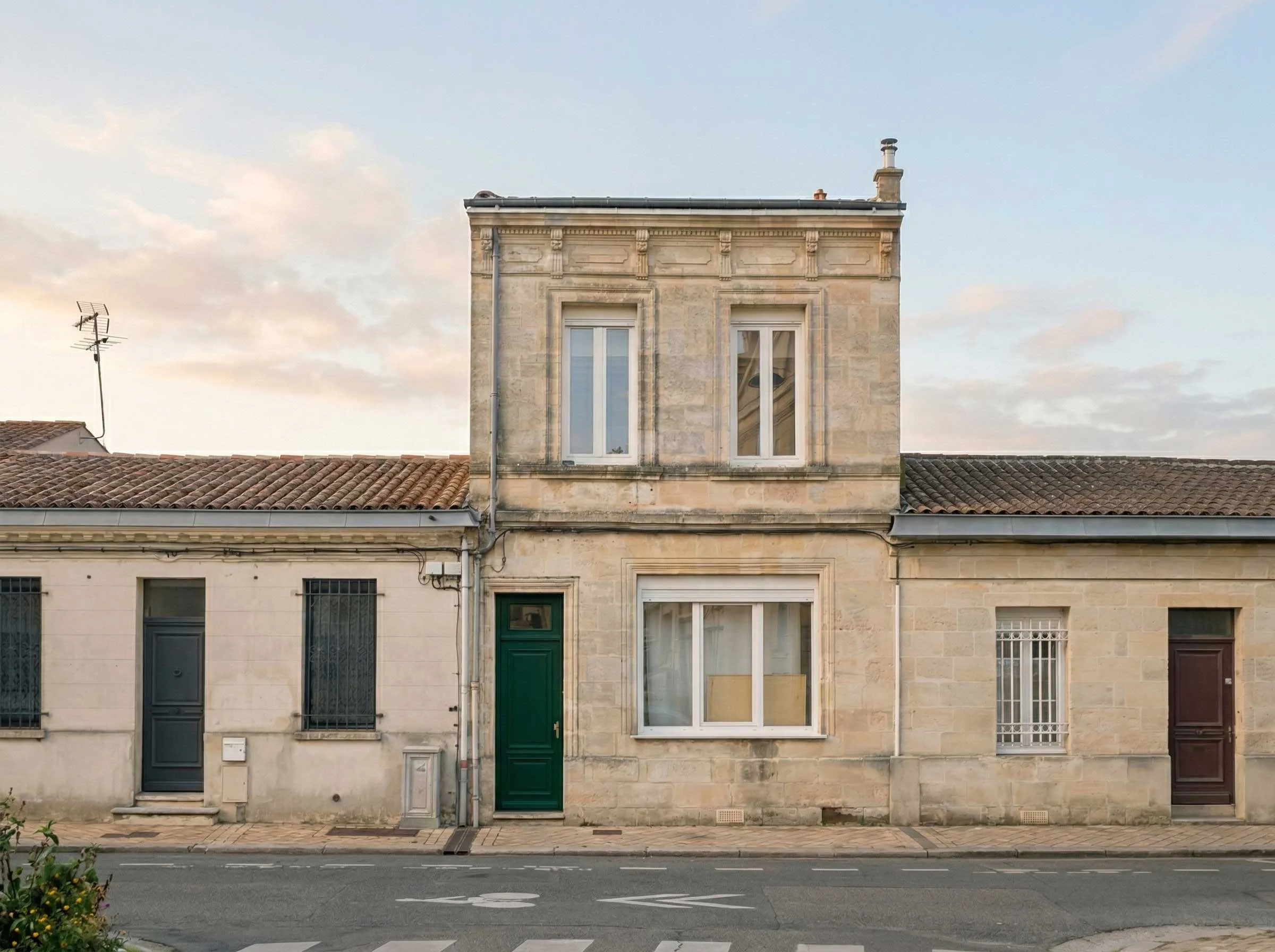 Façade d'immeuble en pierre avec deux fenêtres et une porte en bas. Toit en tuiles avec cheminée et antenne TV. Ciel partiellement nuageux en arrière-plan.