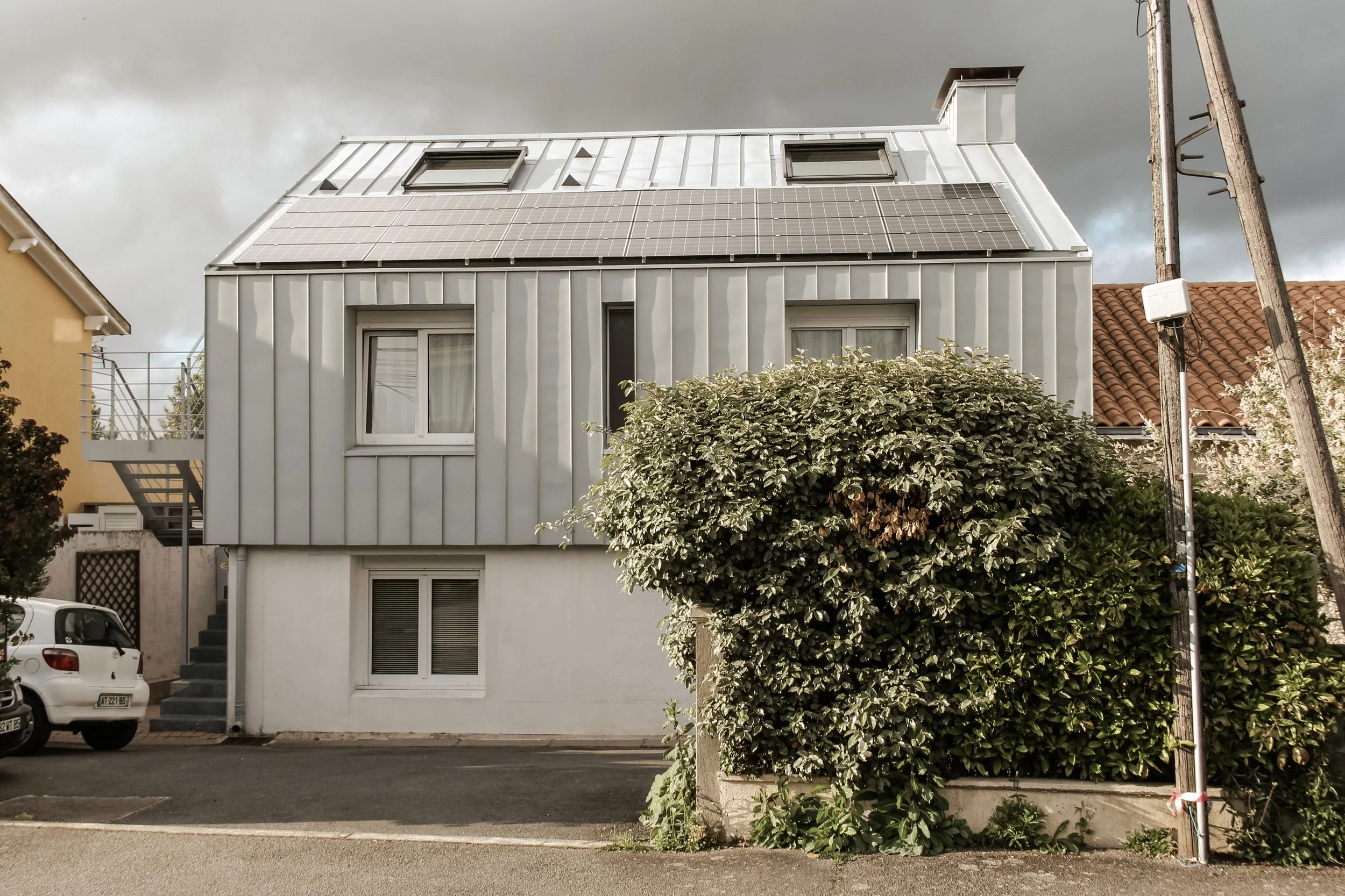 Immeuble blanc avec deux fenêtres et un toit solaire, devant un buisson et une route, avec un ciel nuageux en arrière-plan.