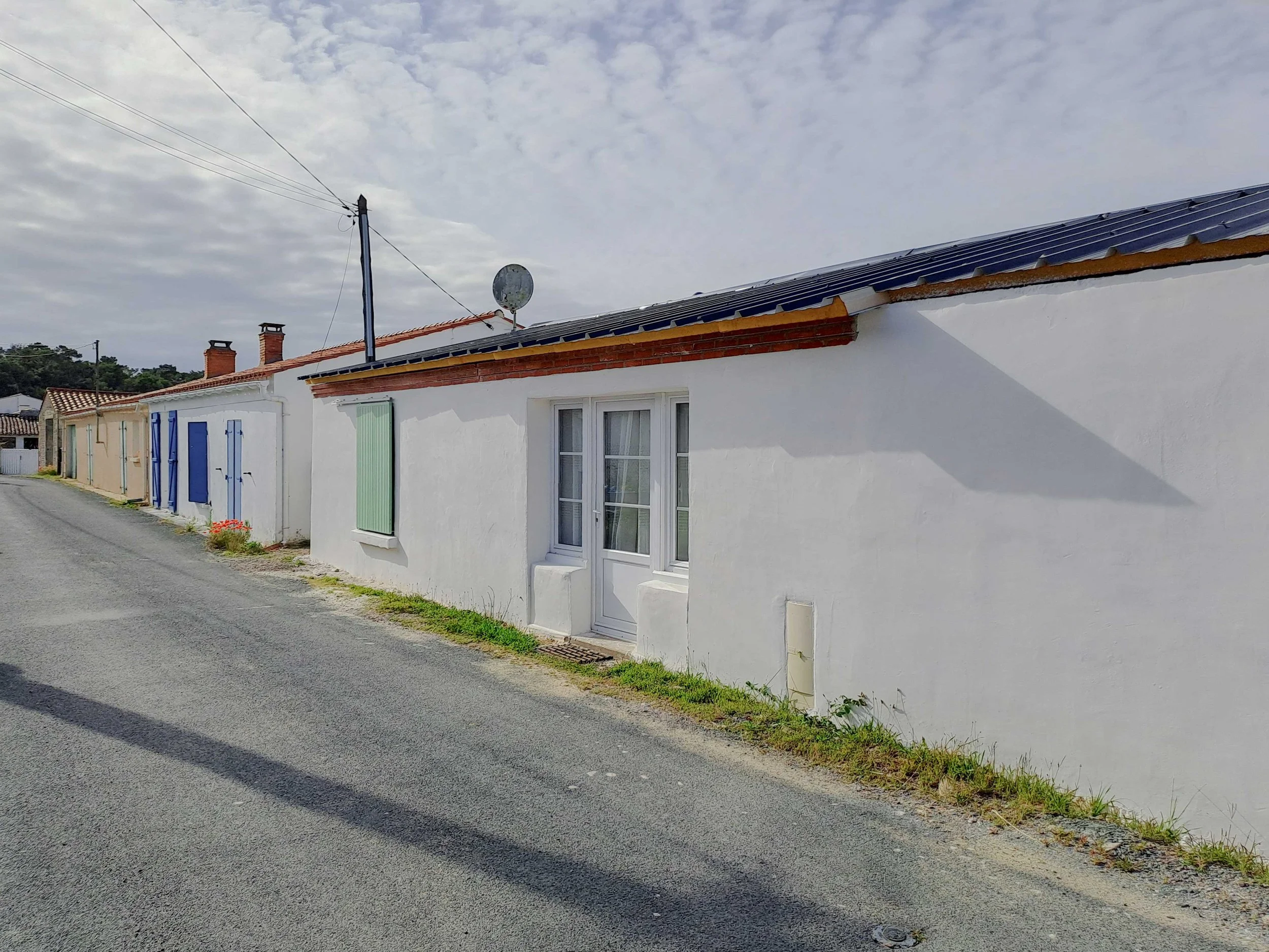 Une maison blanche avec des volets bleus, une fenêtre, une porte en verre, un toit en tuiles, une antenne satellite, un poteau électrique, une rue en pavé et un ciel nuageux.
