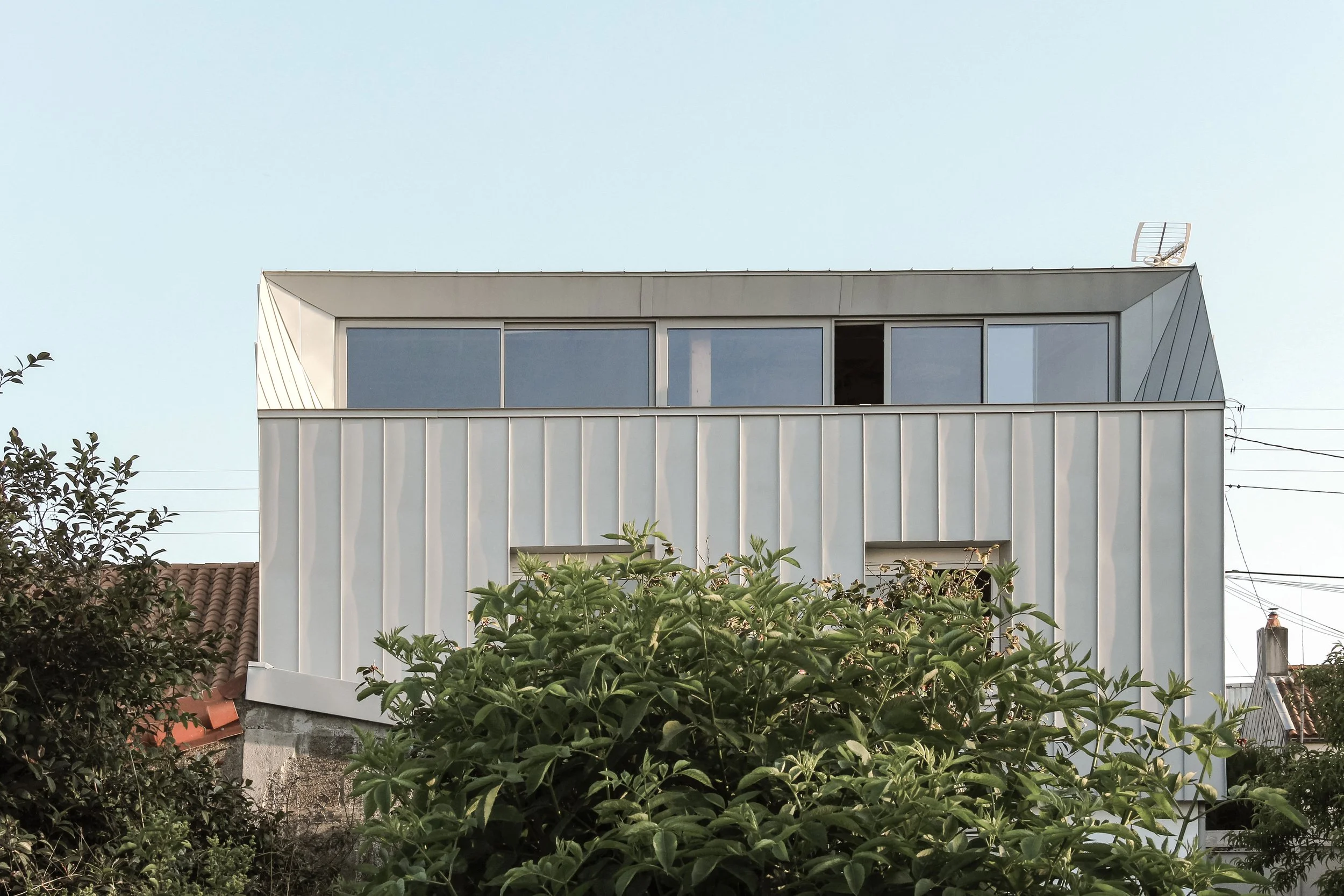 Un bâtiment moderne avec de grandes fenêtres en haut, entouré de végétation au premier plan avec des arbres et des plantes, sous un ciel clair.