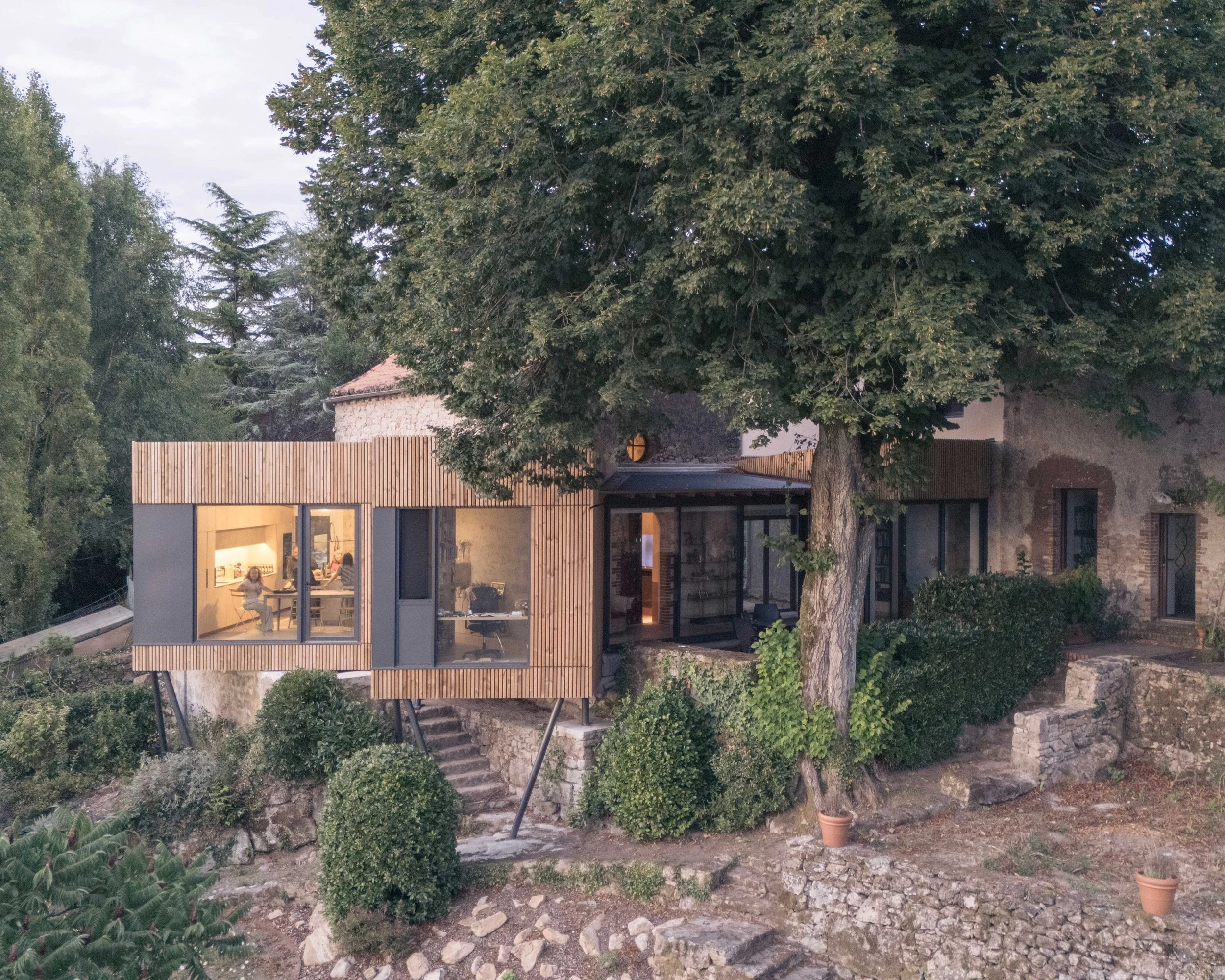 Une maison contemporaine avec grand fenêtre, construite en pierre et bois, entourée d'arbres et de câbles électriques, avec un espace extérieur avec des plantes et des escaliers.