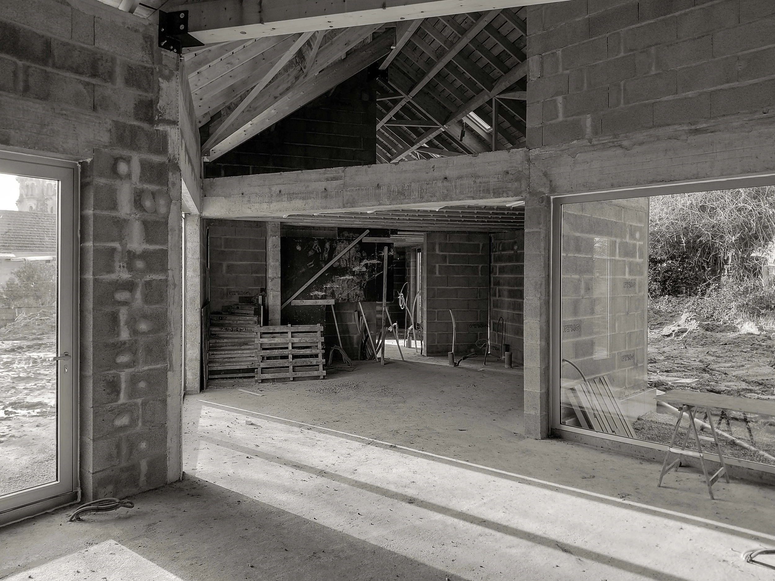 Intérieur d'une maison en construction avec murs en briques, plans de travail en béton, poutres en bois apparentes sur le toit et grandes fenêtres sans vitrage laissant entrer la lumière naturelle.
