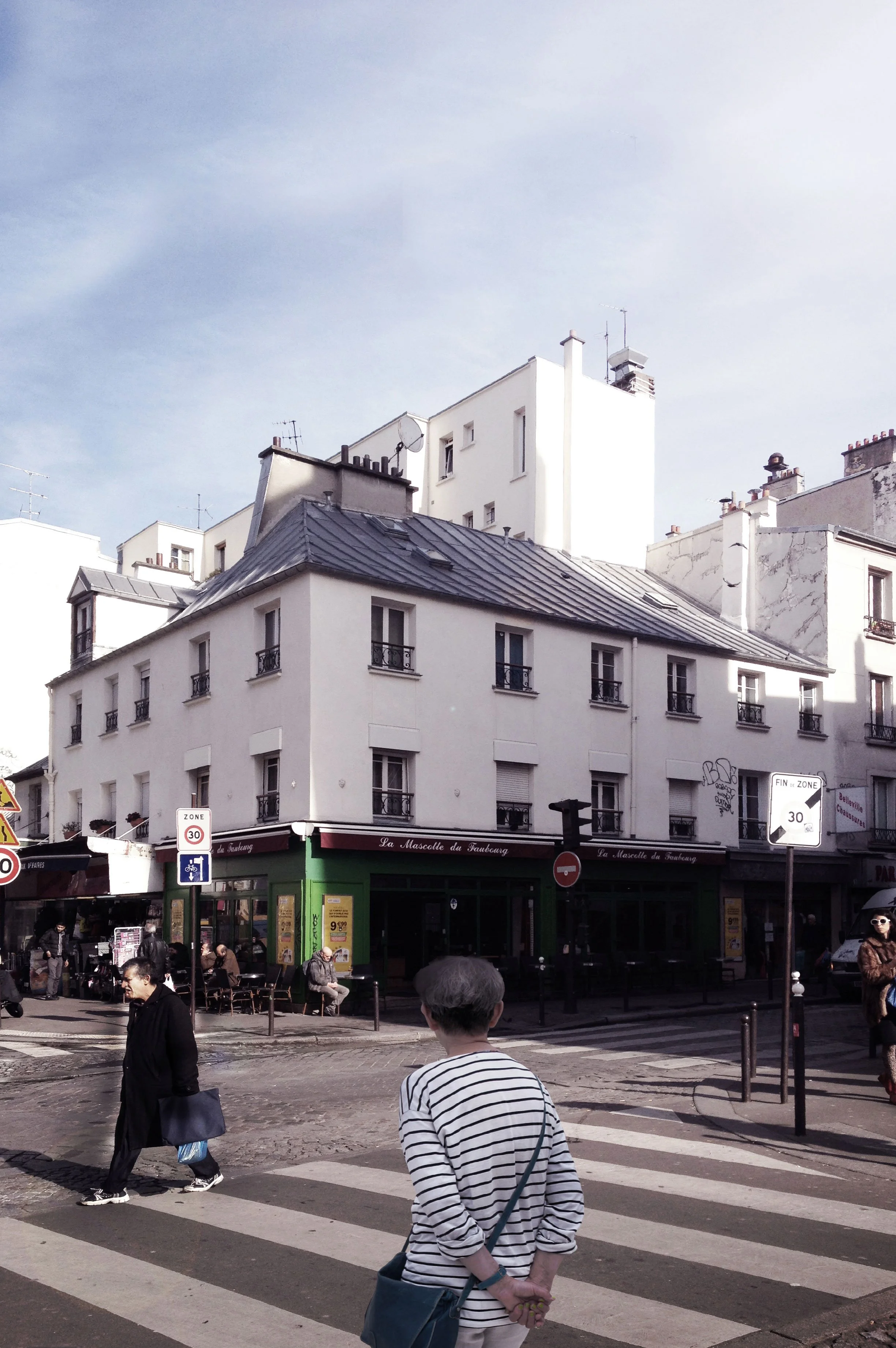 Vue urbaine avec des bâtiments blancs à plusieurs étages, un restaurant au rez-de-chaussée, des piétons traversant une rue avec des panneaux de signalisation, une zone limitée à 30 km/h, et un ciel clair.