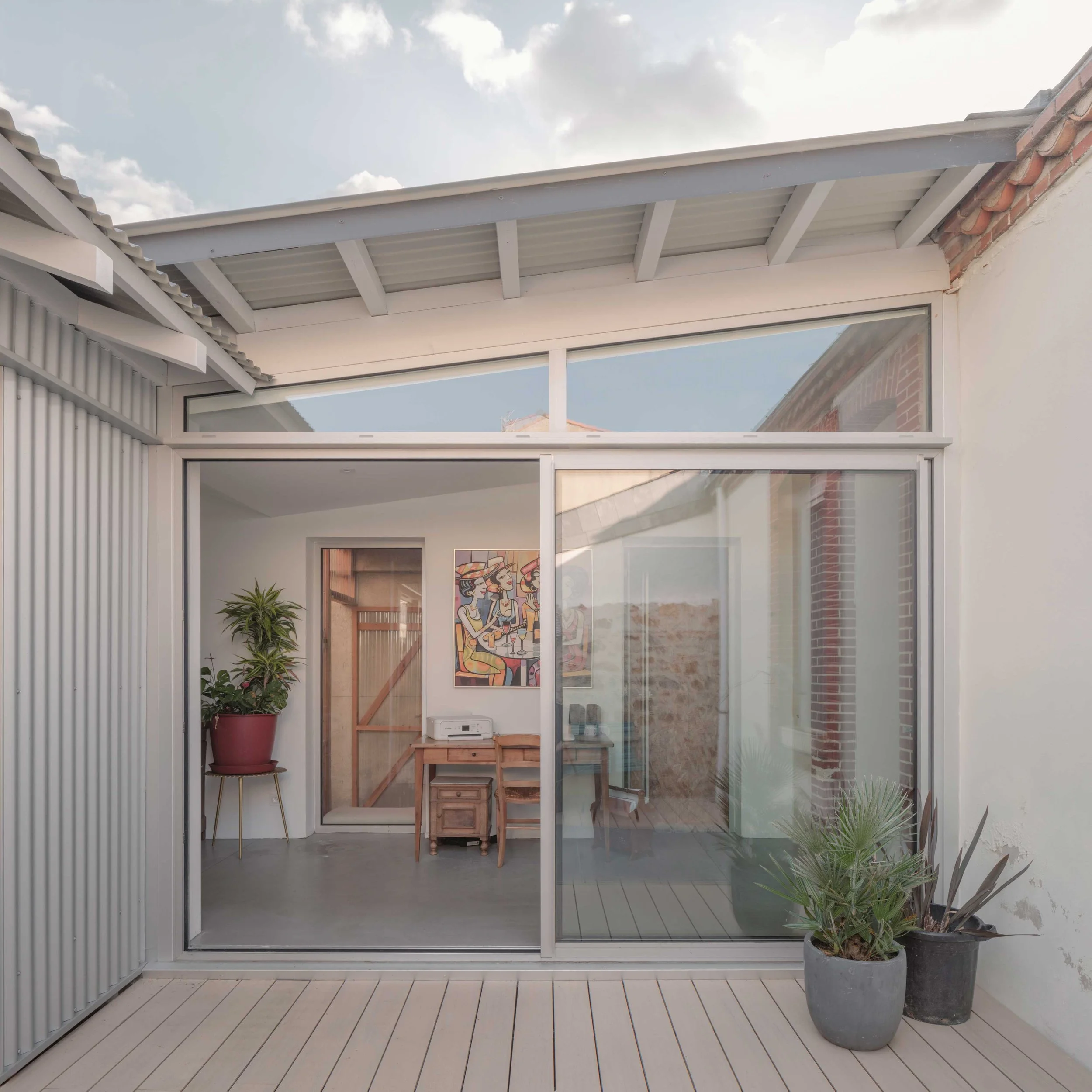 Une véranda moderne avec de grandes fenêtres en verre, un sol en bois clair, et des plantes d'intérieur, avec une peinture murale colorée visible à l'intérieur.