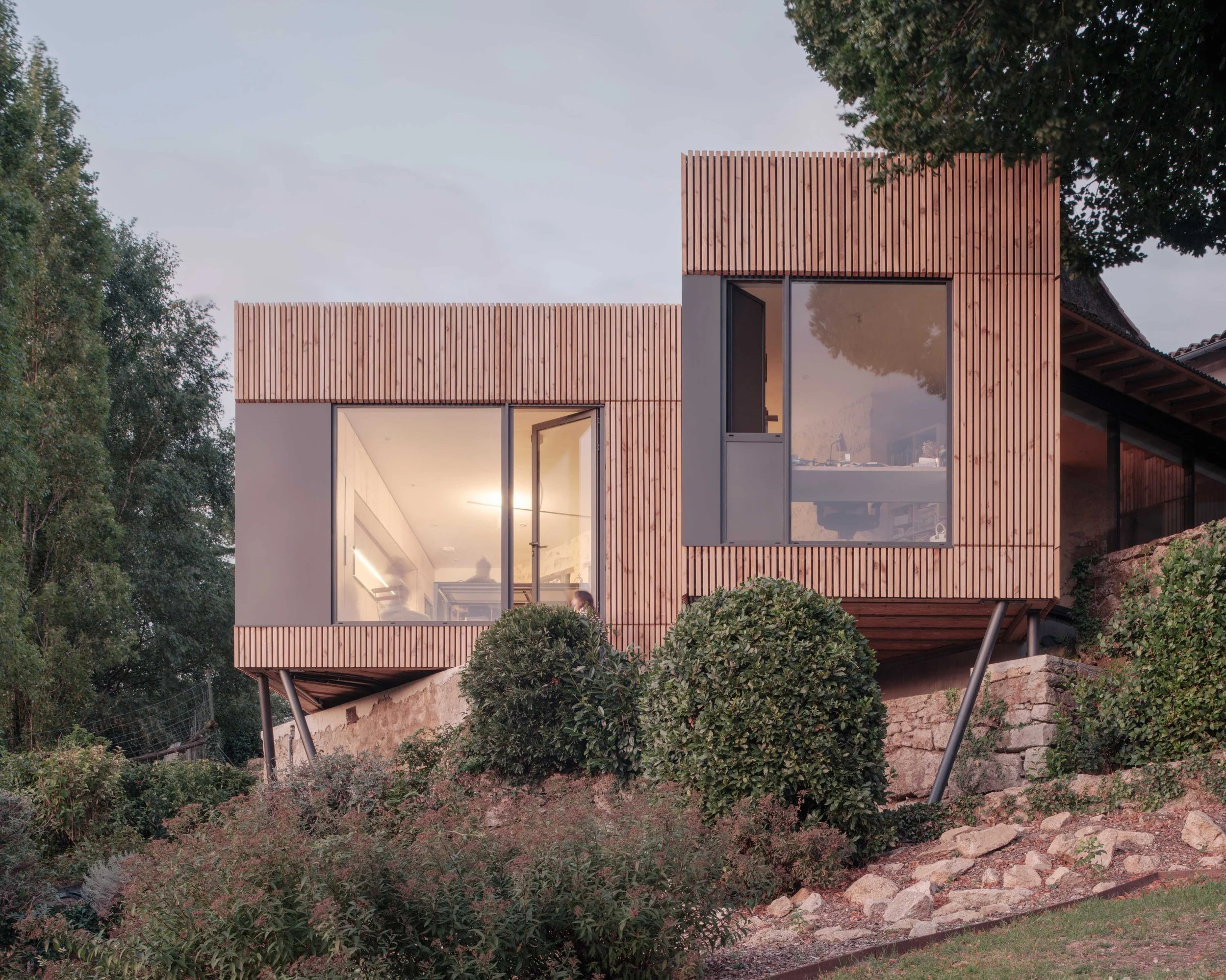 Facade moderne en bois avec grandes fenêtres dans un jardin avec des arbres et des buissons.
