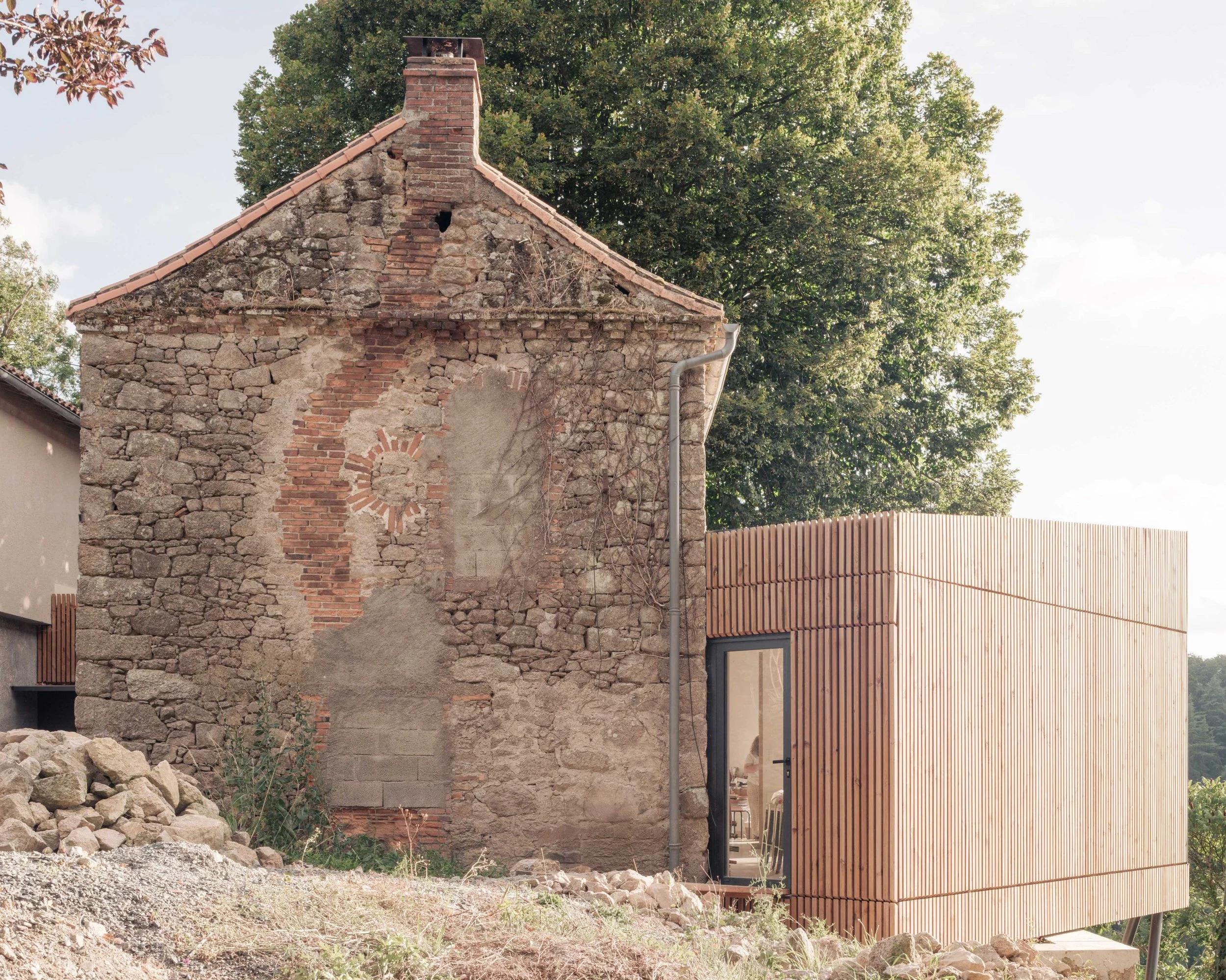 Une ancienne bâtisse en pierre partiellement en ruine avec une construction moderne en bois à droite. Un arbre feuillu en arrière-plan et un ciel partiellement nuageux.