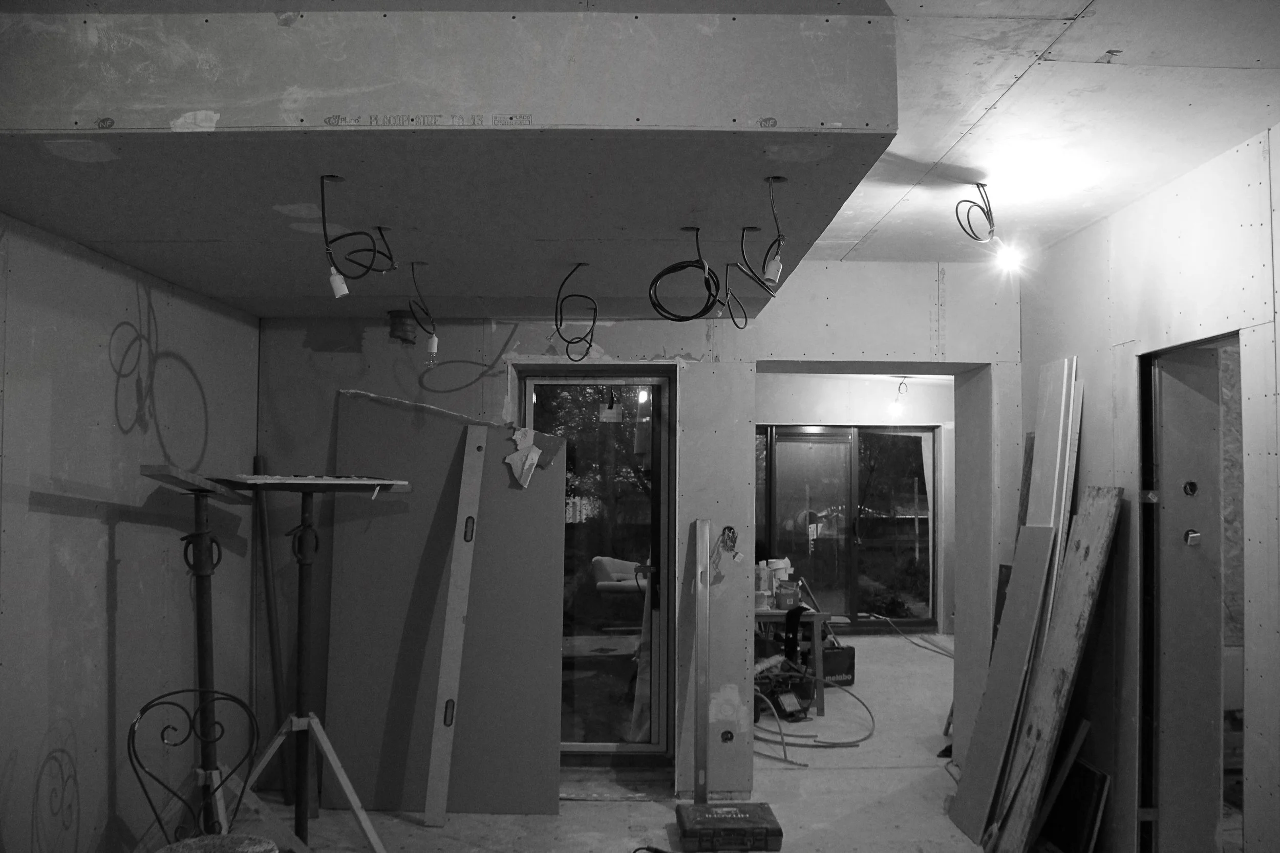 Une pièce en cours de construction ou de rénovation, avec des murs et un plafond en bois ou en plaque de plâtre, des câbles électriques suspendus, des outils et des matériaux dispersés, sans meubles ou décorations.
