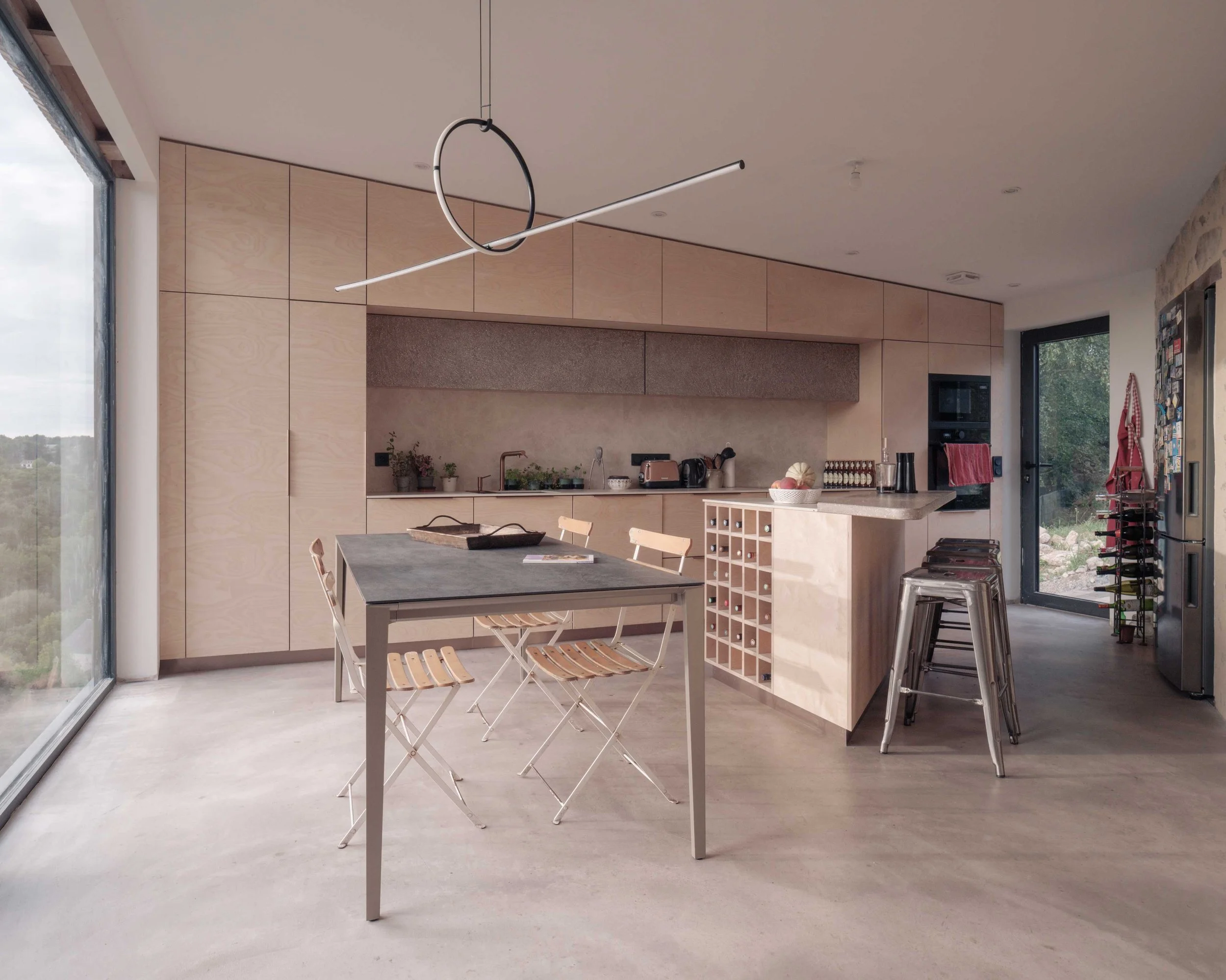 Une cuisine moderne avec un plan de travail, des tabourets en métal, une table avec des chaises pliantes en bois, une grande fenêtre laissant entrer la lumière naturelle, et un plafond avec une lampe design suspendue.