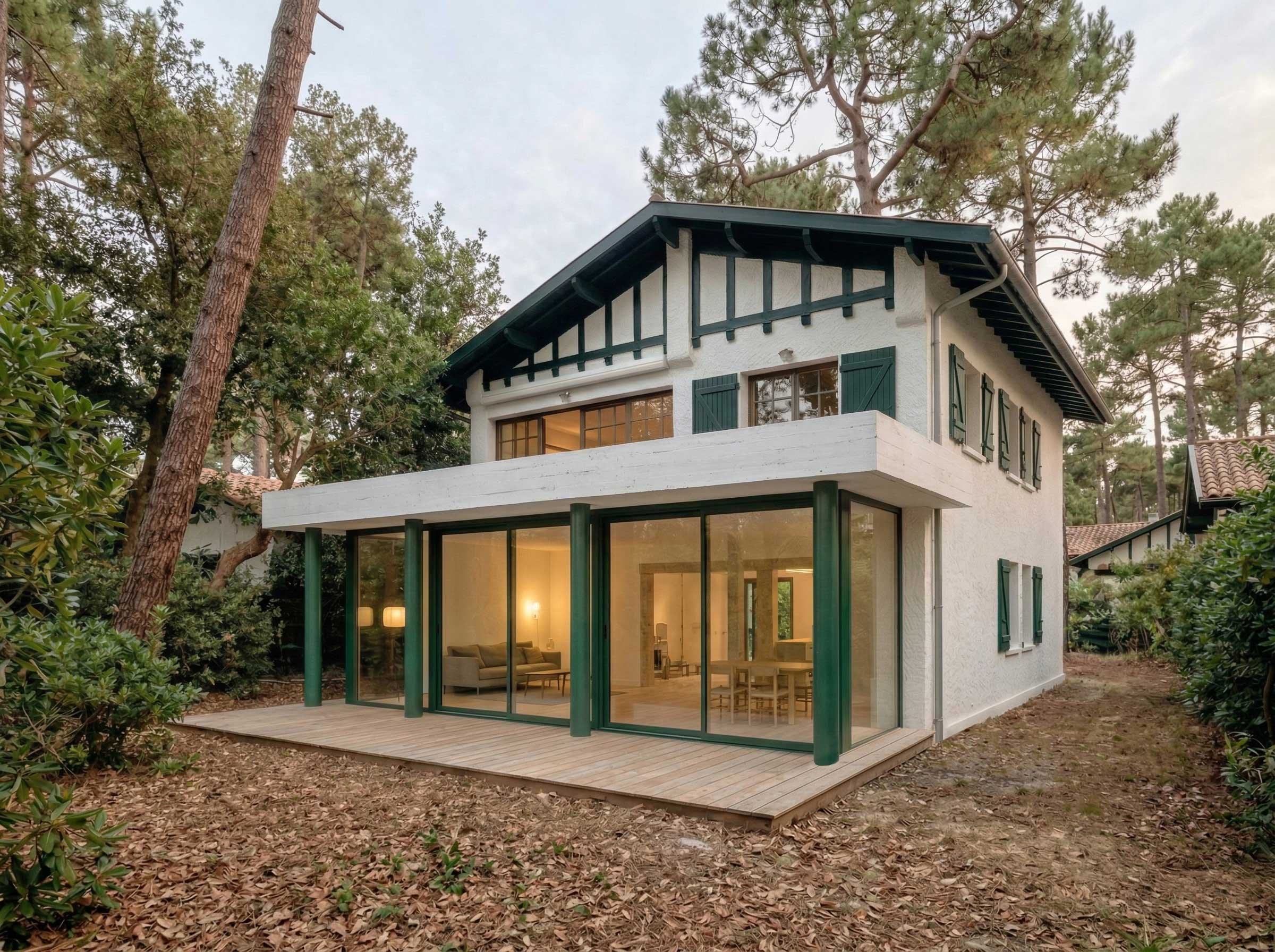 Une maison à deux étages avec une grande baie vitrée au rez-de-chaussée, entourée d'arbres et de végétation, avec un patio en bois.