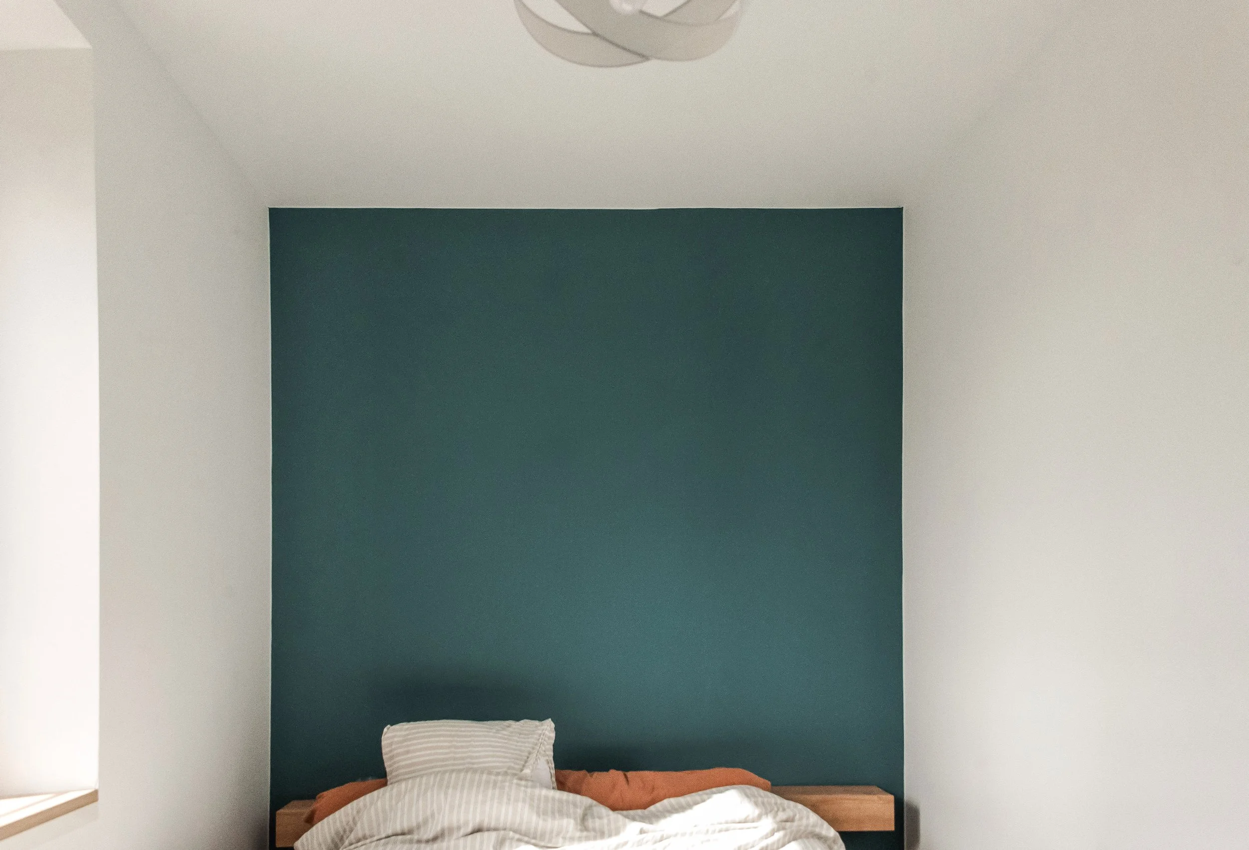 Photo d'une chambre avec un mur bleu derrière le lit, un drap beige et une couette orange, une lampe design au plafond et une fenêtre à gauche.