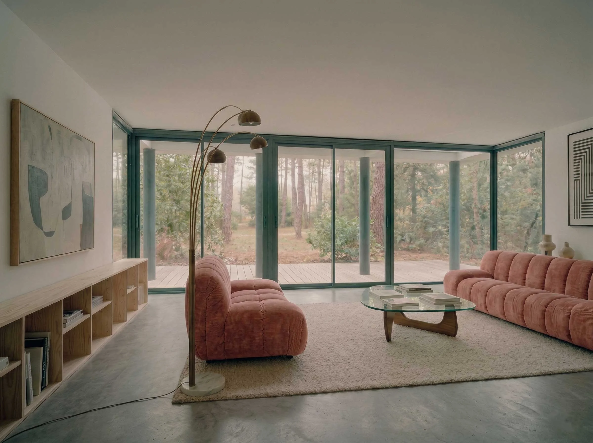Salon moderne avec grandes fenêtres, canapé en velours rose, table en verre, étagère en bois avec livres, peinture abstraite au mur, lampadaire arc en métal et lampe sur la table.