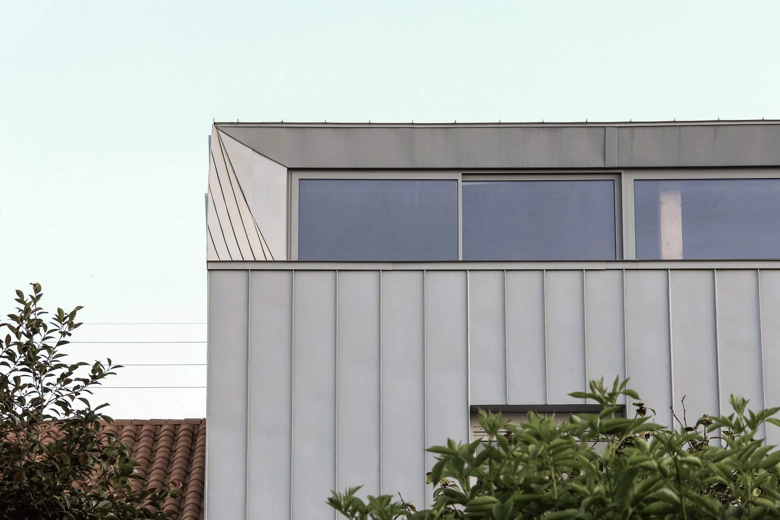 Vue partielle d'un immeuble moderne avec des fenêtres en verre et une façade en aluminium, partiellement obstruée par des arbres devant.