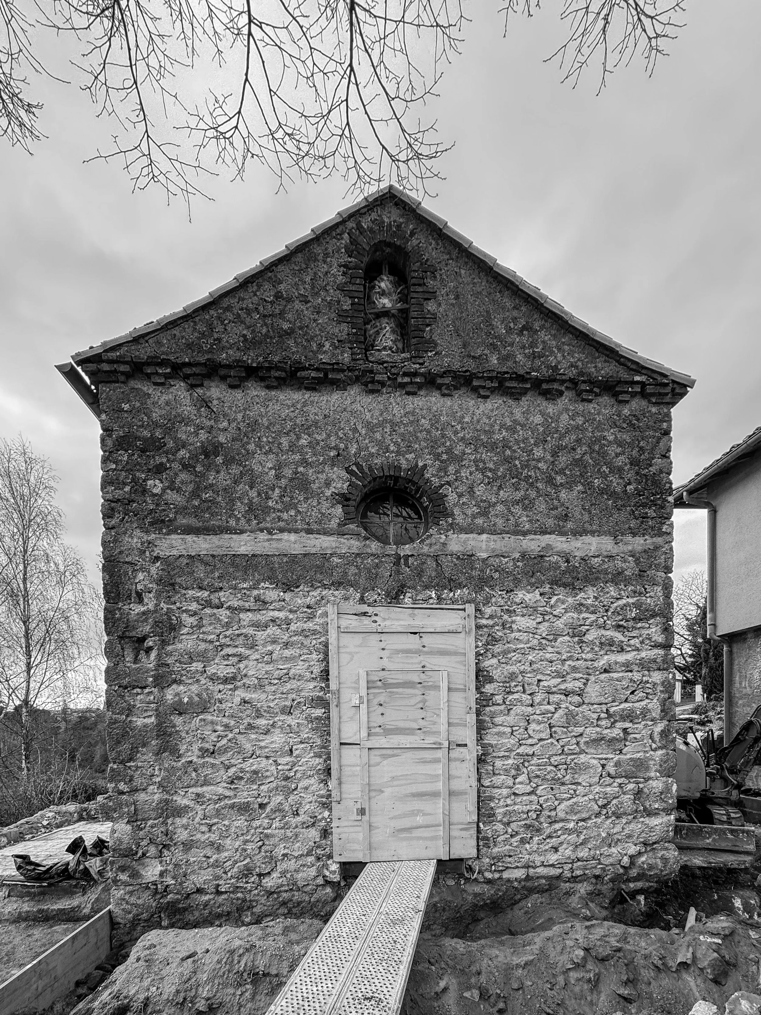 Une vieille bâtisse en pierre avec une fenêtre à arc en haut, un panneau en bois devant une porte, une rampe métallique, et des branches d'arbres sans feuilles en arrière-plan, sous un ciel nuageux.