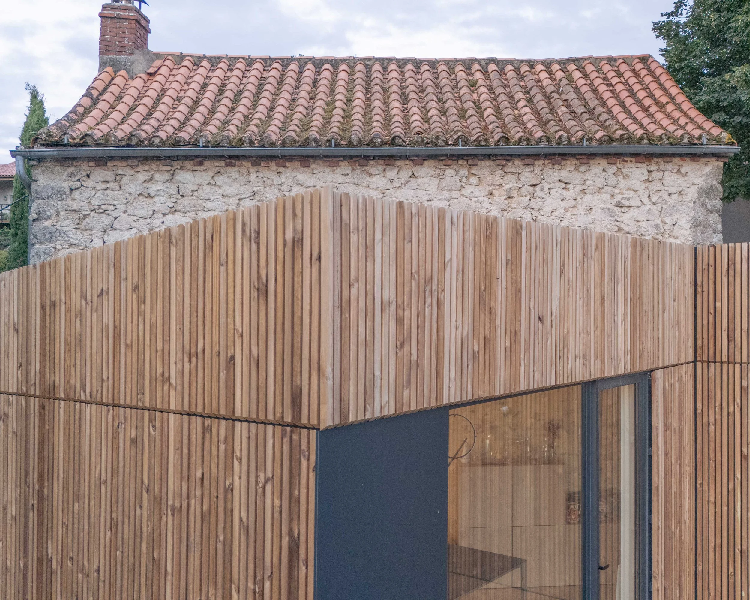 Façade d'une maison en pierre avec un toit en tuiles anciennes et un mur en bois devant.