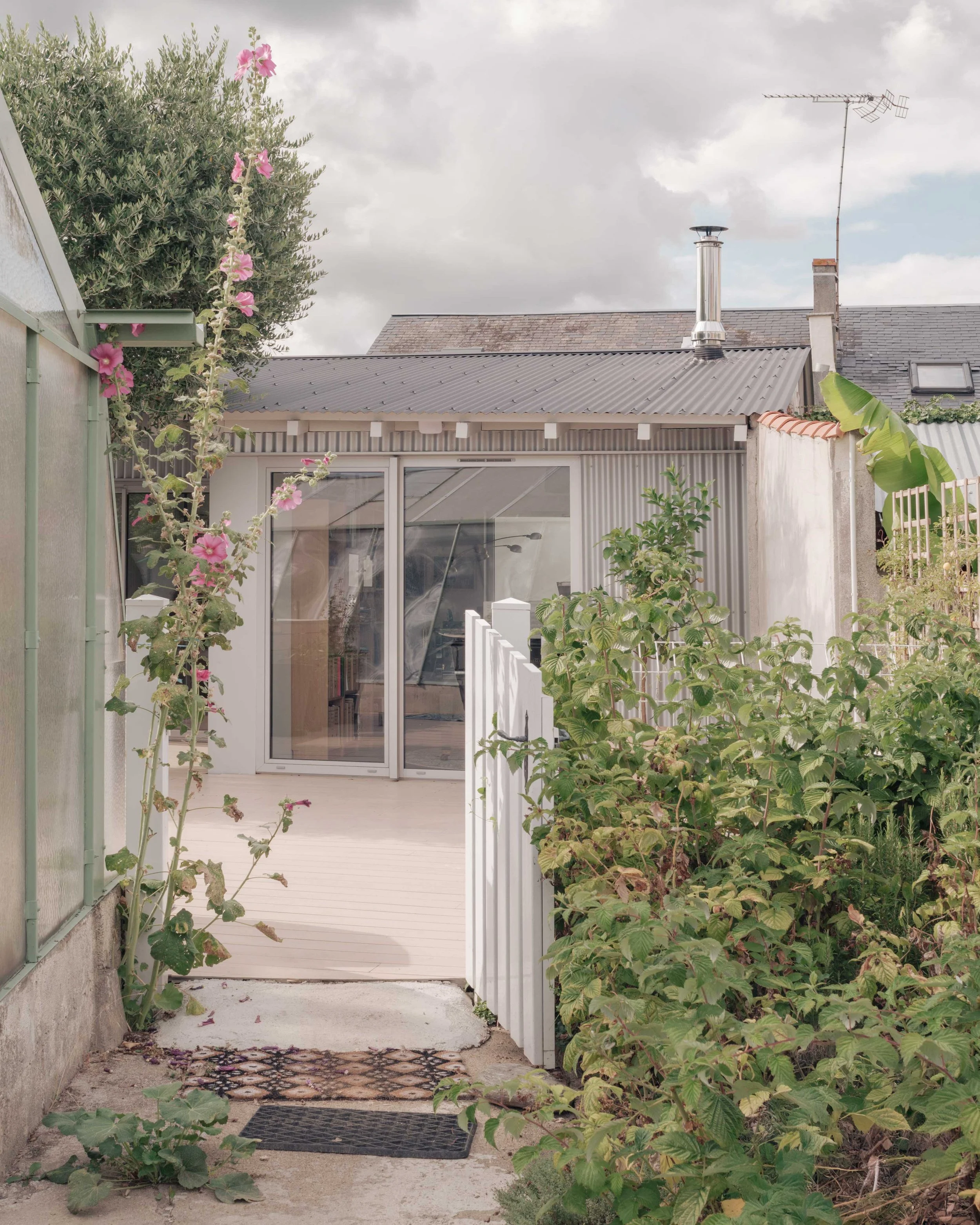 Une cour extérieure avec une porte-fenêtre en verre, entourée de plantes et de végétation, un tapis de porte en caoutchouc noir et un matelas à l'entrée.