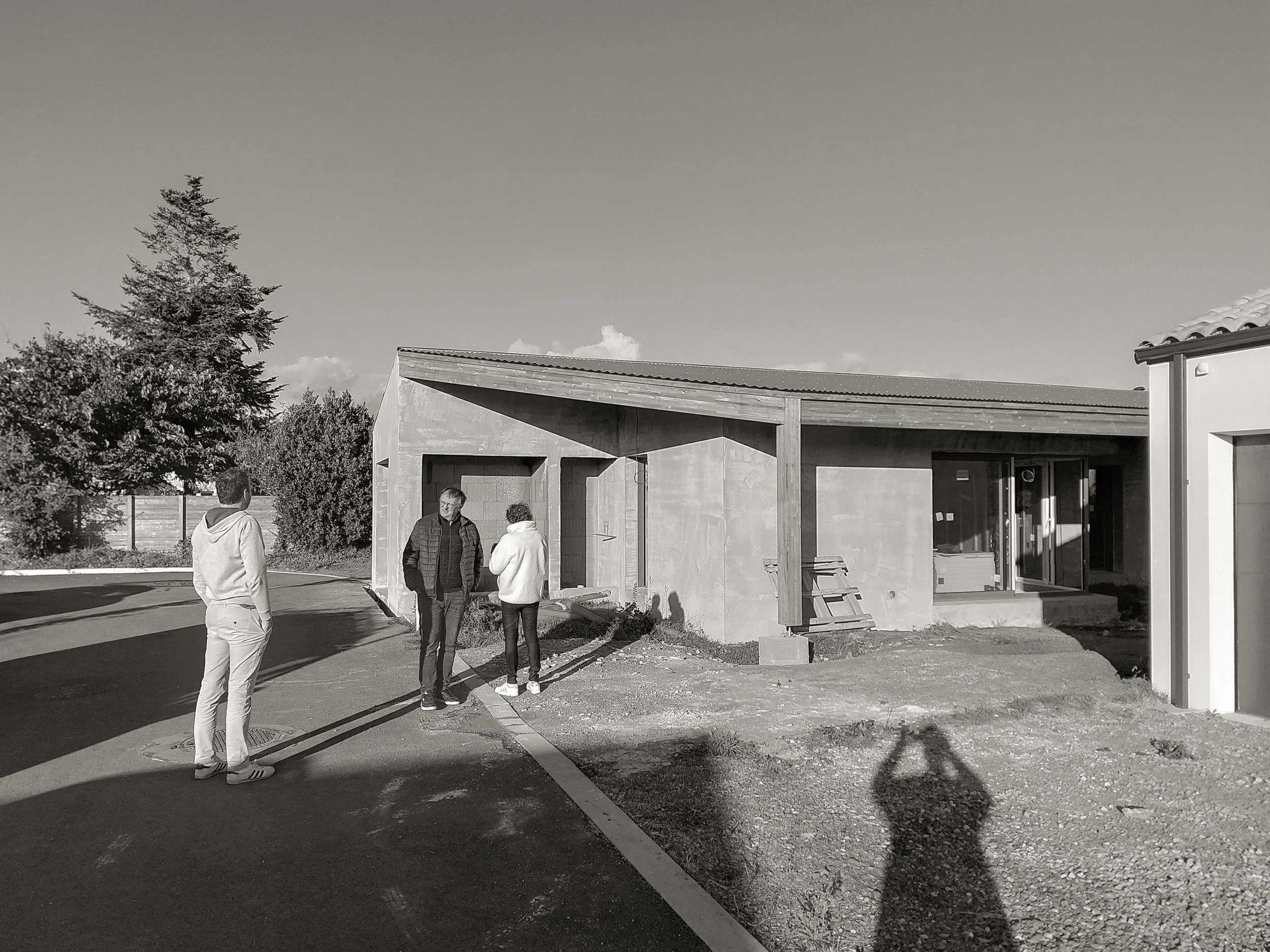 Trois personnes discutent devant un bâtiment en construction ou récemment construit, avec un toit en tuiles et des murs en béton, dans un environnement extérieur avec des arbres et un ciel clair.