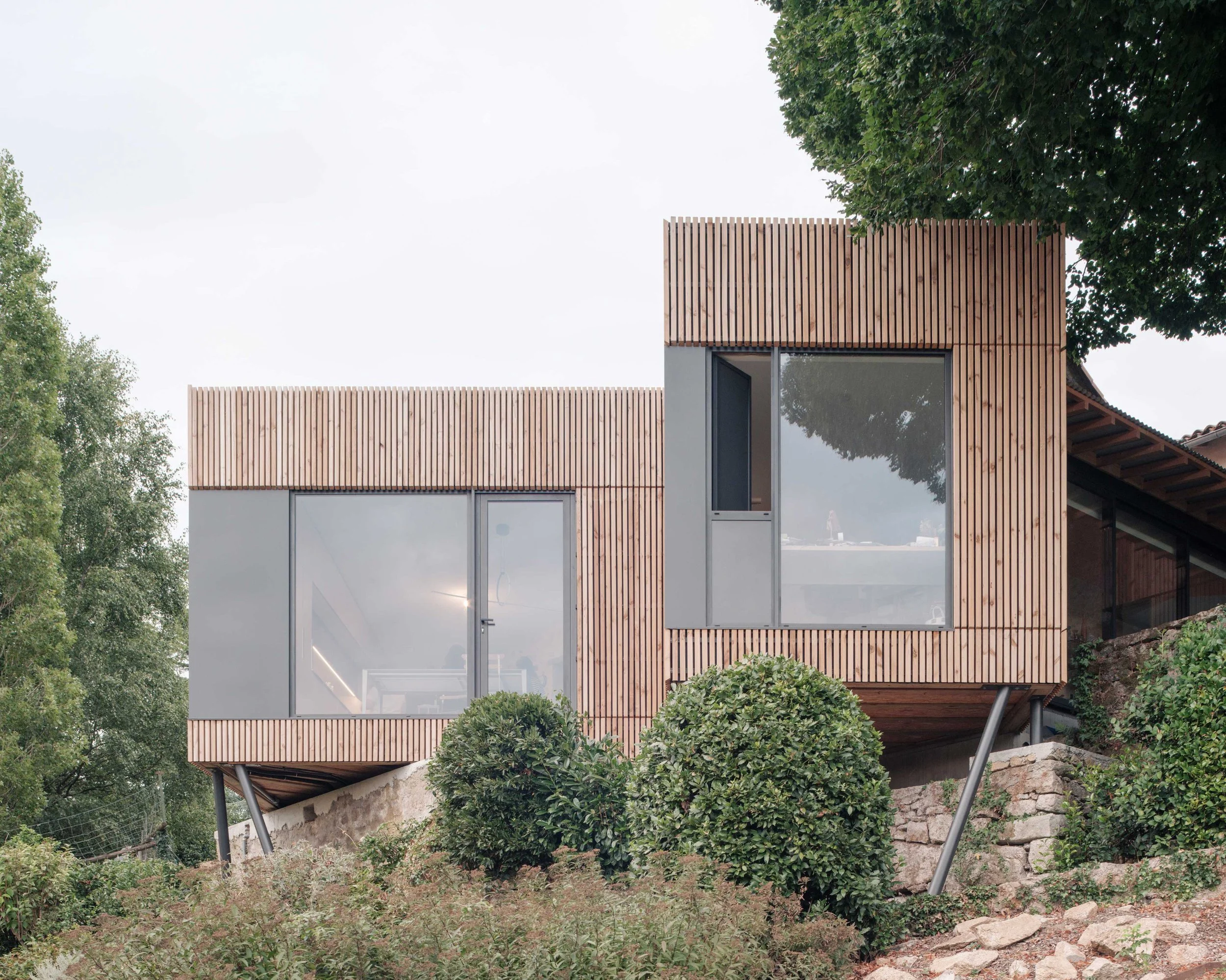 Une façade de bâtiment moderne en bois et en verre entourée de verdure, avec des arbres et des buissons.