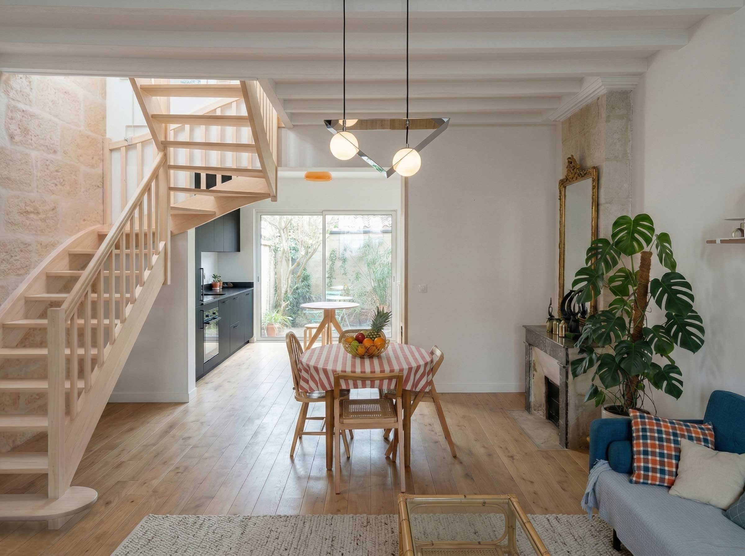 Un intérieur lumineux avec escaliers en bois, table à manger ronde avec nappe à rayures rouges et blanches, cuisine noire en arrière-plan, grand miroir doré sur le mur, plante verte à droite, fauteuil bleu avec coussin à carreaux à gauche.