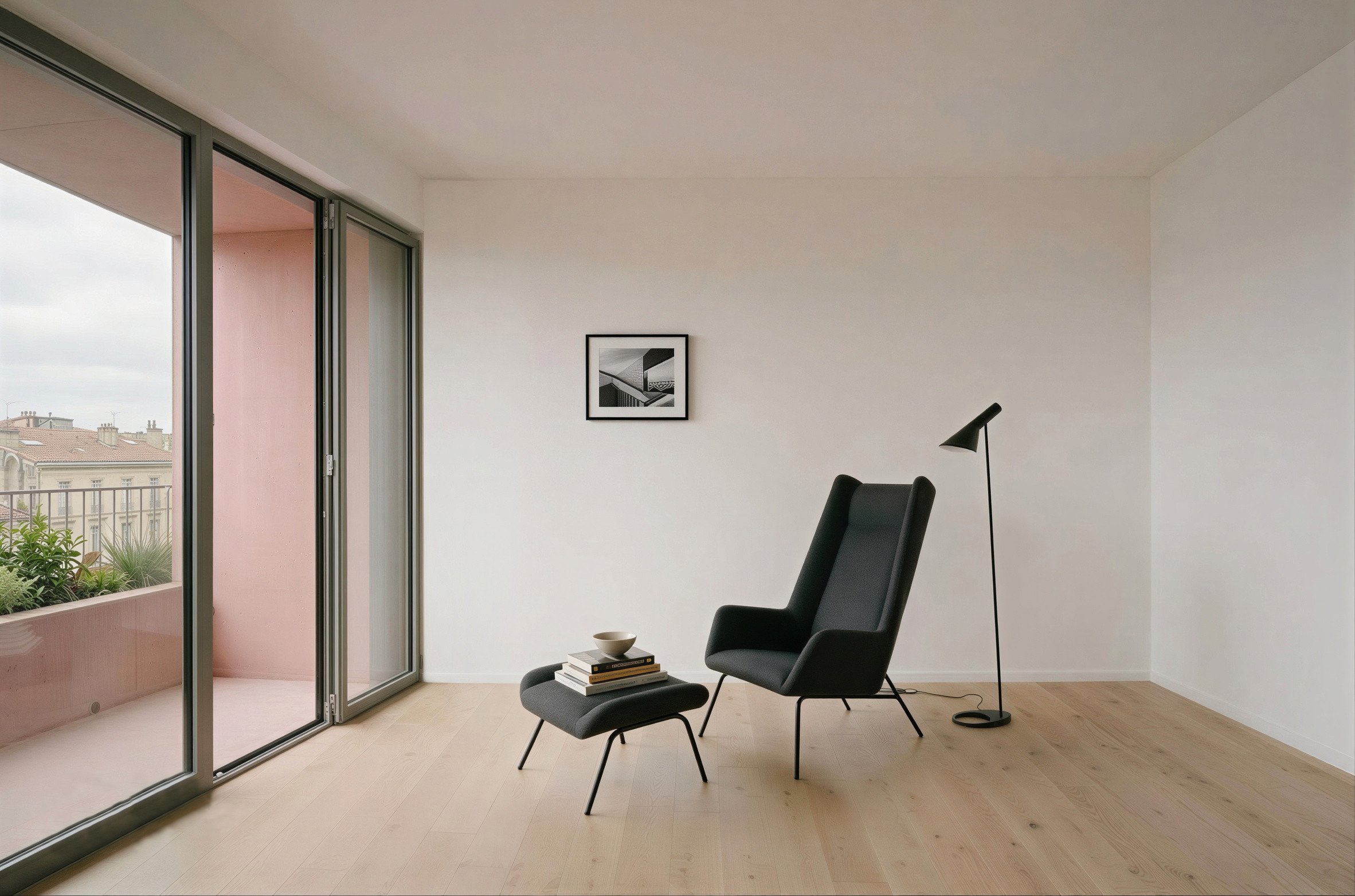 Intérieur d'une pièce moderne avec un fauteuil, une table avec des livres et une lampe d'appoint, et une grande fenêtre ouvrant sur un balcon avec vue sur le ciel et les toits des bâtiments voisins.
