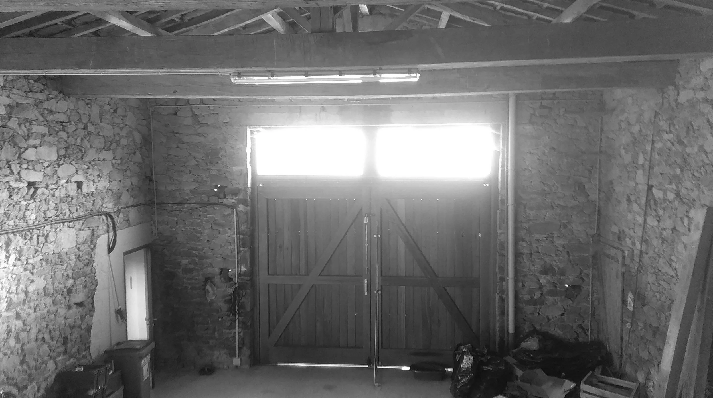 Un garage en construction avec des murs en pierre et une porte en bois avec des carreaux en verre au sommet, éclairé par une lumière naturelle venant d'une fenêtre.