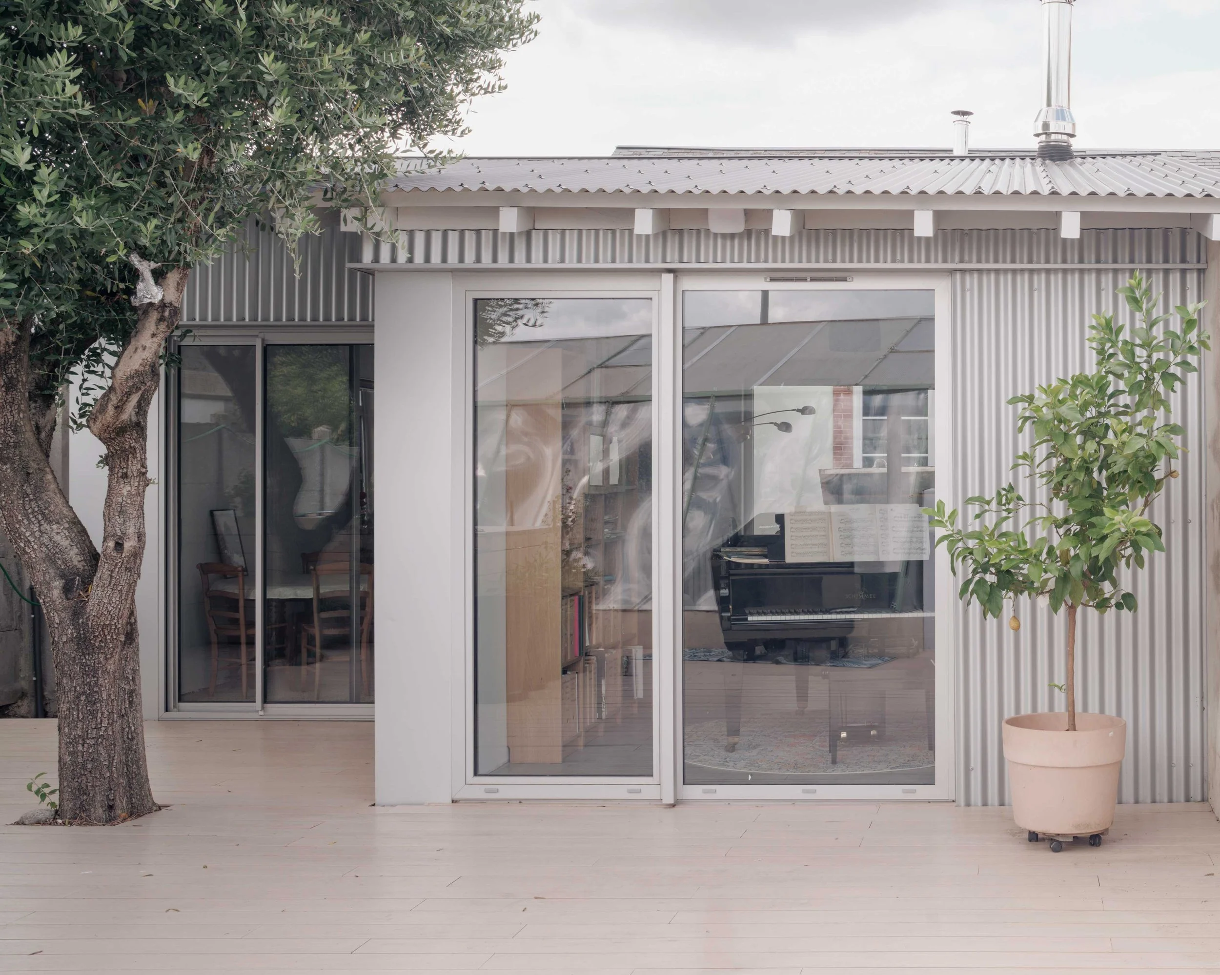 Une maison moderne avec un mur en métal ondulé, un panneau en verre laissant voir l'intérieur, une porte-fenêtre, un arbre et un pot en terre cuite avec une plante à l'extérieur.