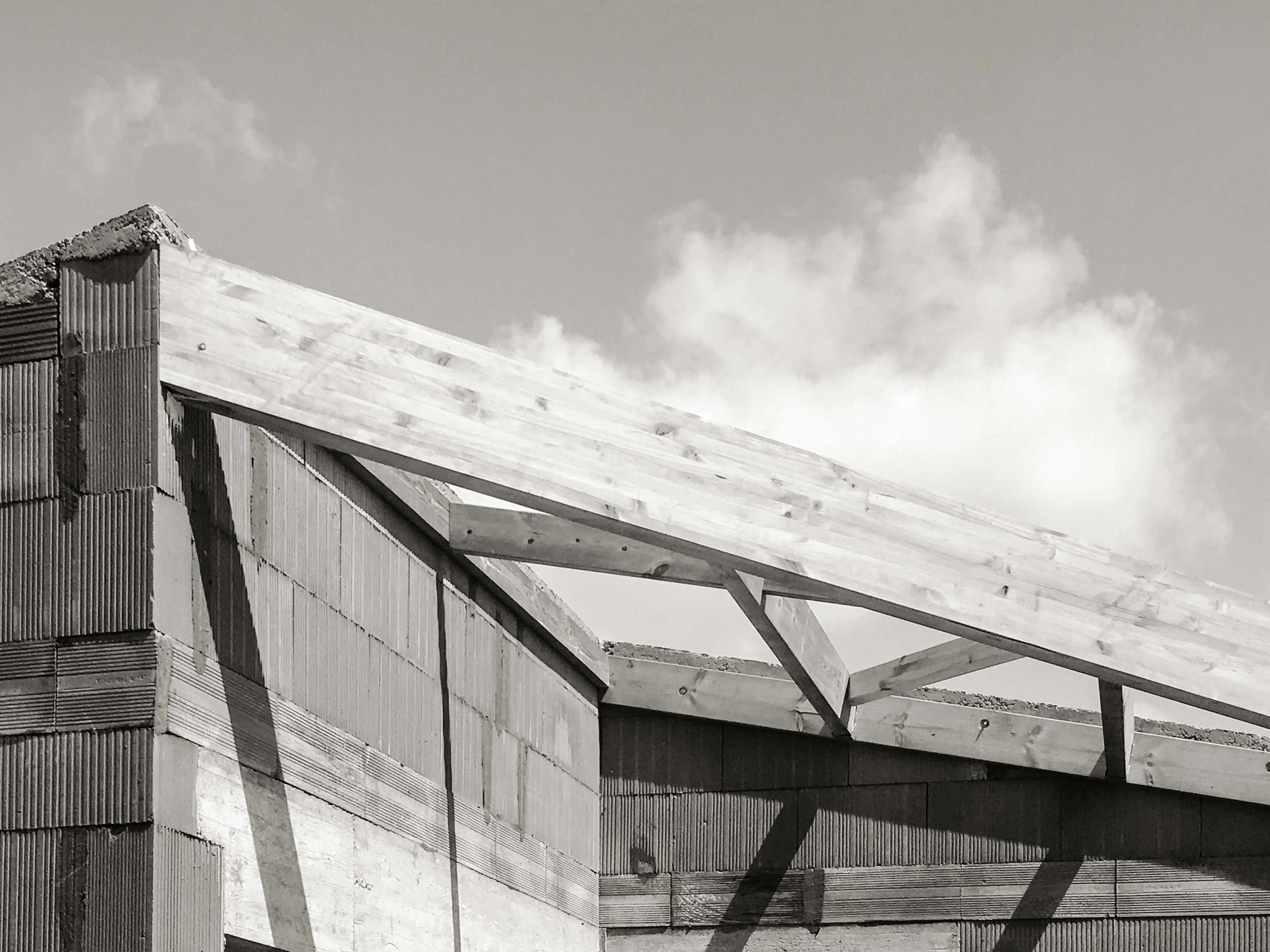 Photo en noir et blanc d'une construction en cours, avec une structure en bois et en métal, sous un ciel nuageux.