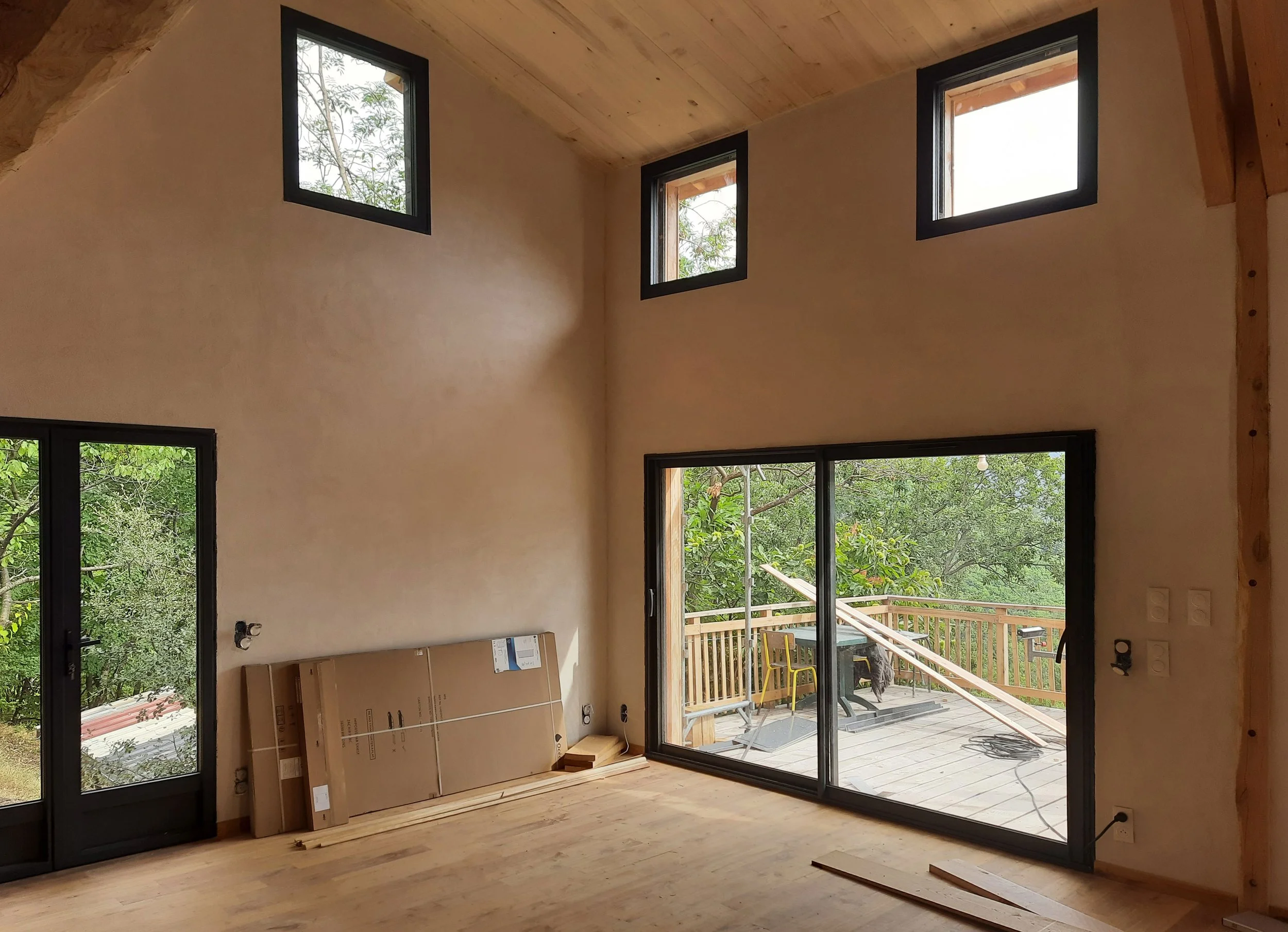 Intérieur d'une pièce en construction avec fenêtres et terrasses en bois, vue sur jardin verdoyant.