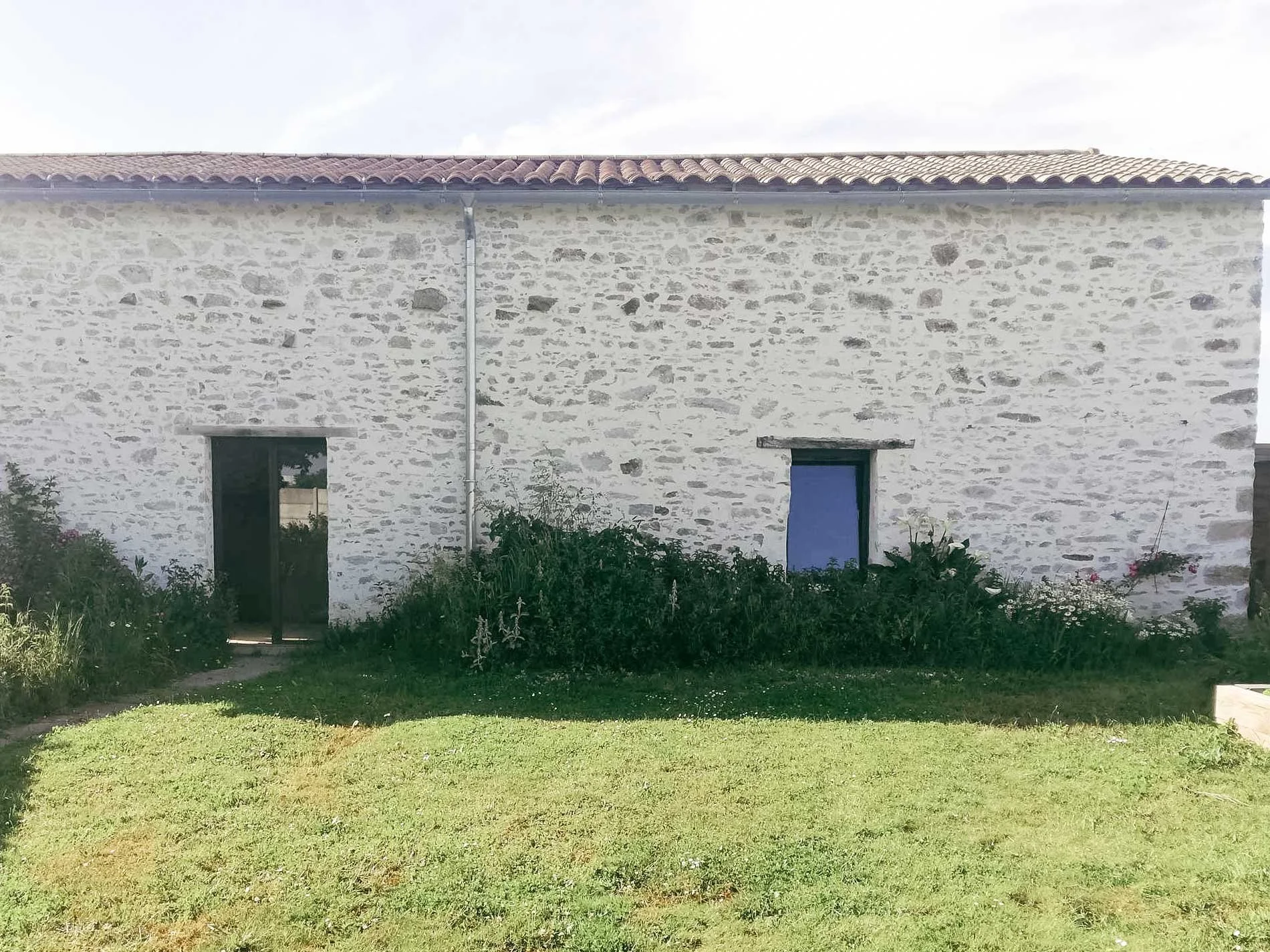 Habitation en pierre avec deux fenêtres et une porte, entourée d'une pelouse verte et de plantes, ciel clair.