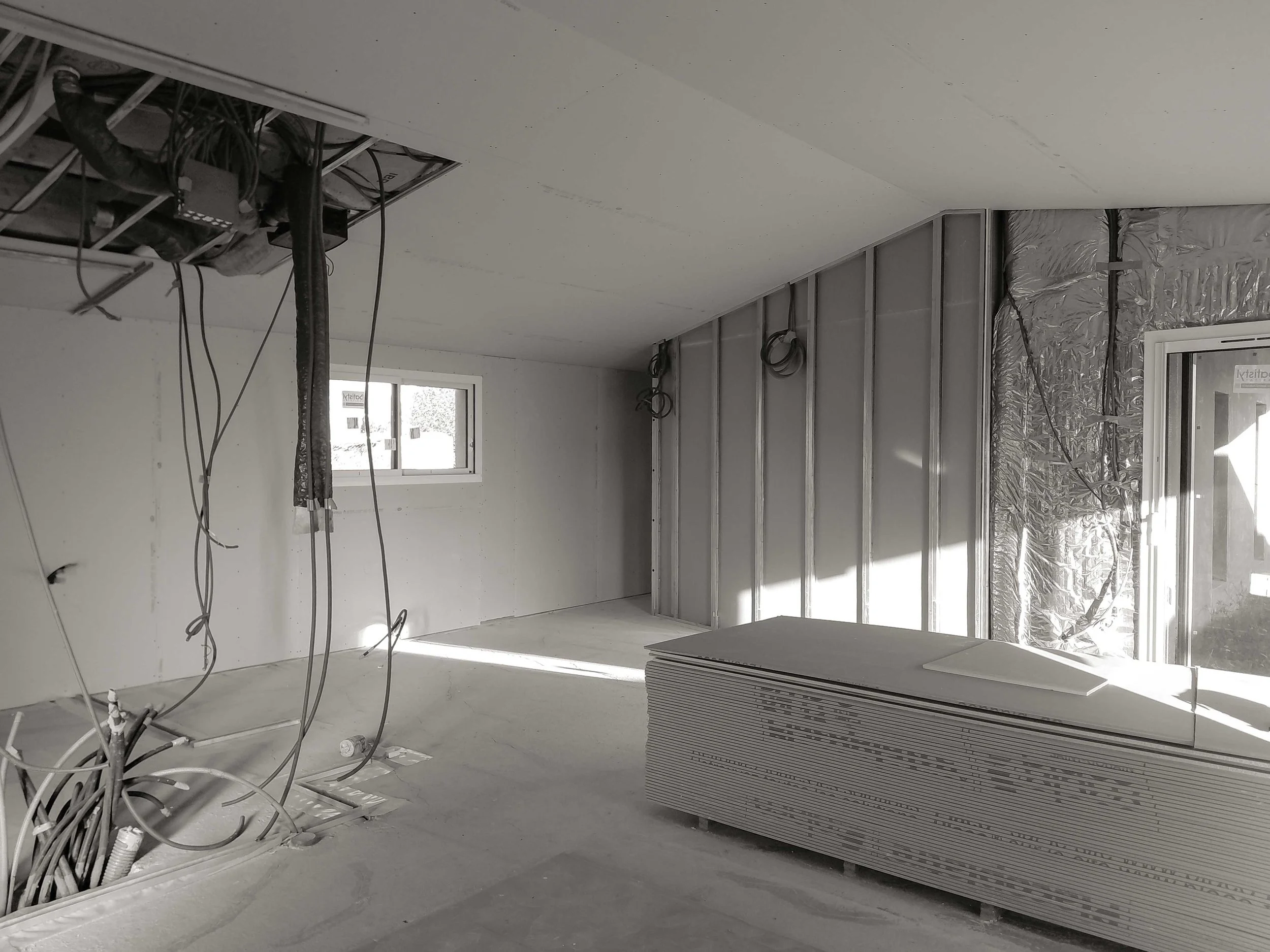 Une pièce en construction avec des murs et un plafond en plâtre non finis, des câbles électriques suspendus, une fenêtre laissant entrer de la lumière naturelle, et un plancher en béton. Des plaques de plâtre stockées sont empilées contre un mur.