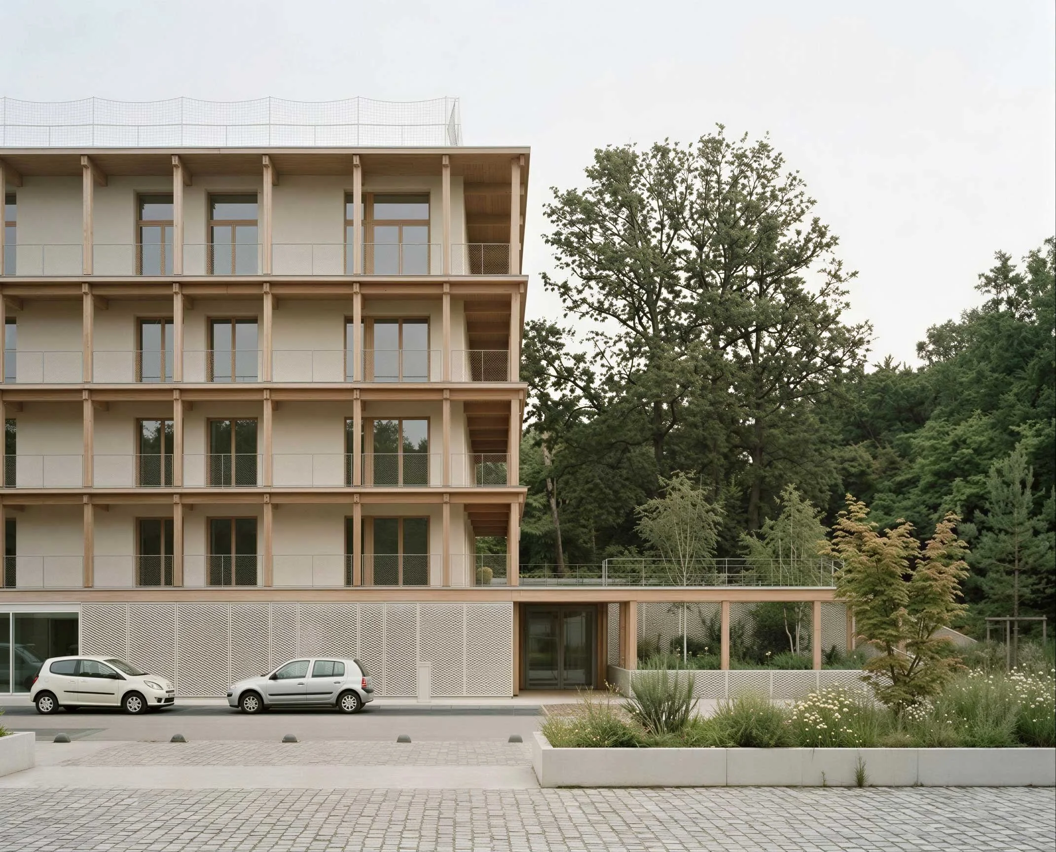 Façade d'un bâtiment moderne avec plusieurs étages, murs beige, fenêtres avec cadres en bois, et balcons, entouré d'arbres et d'arbustes, avec une zone de stationnement avec deux voitures en premier plan.