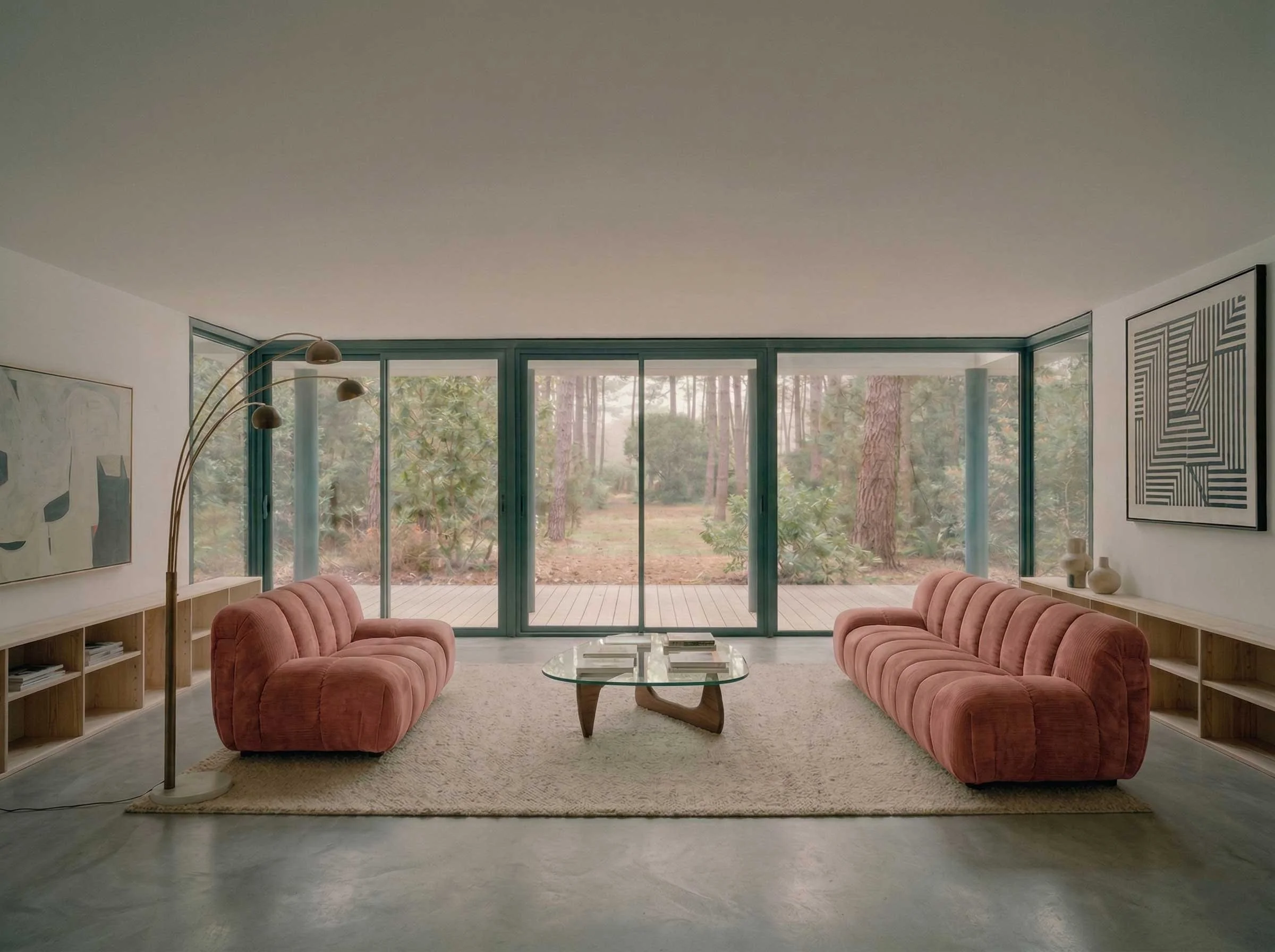 Un salon moderne avec deux canapé en velours rose, une table en verre, et de grandes fenêtres donnant sur une forêt.