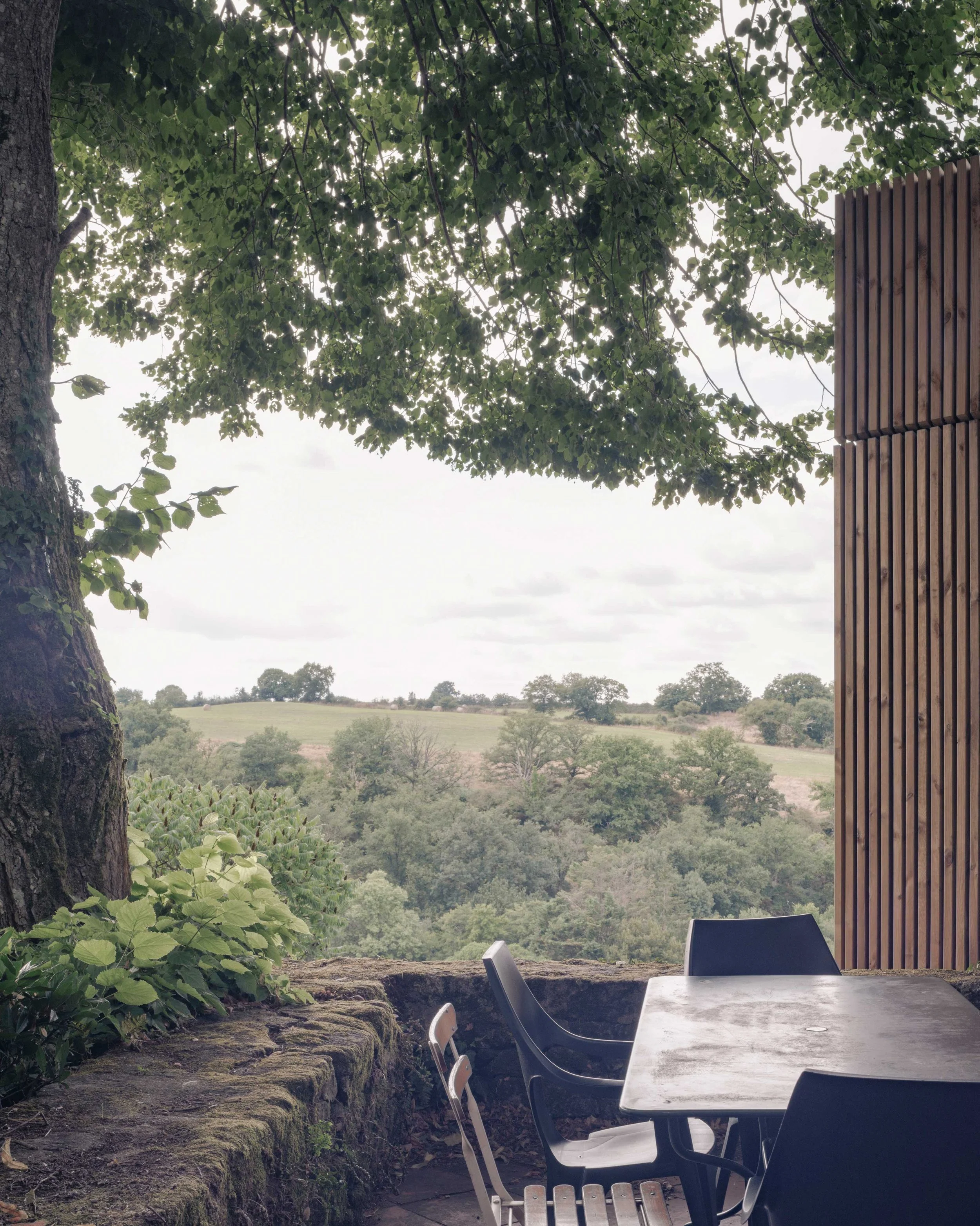 Une terrasse avec une table en pierre et des chaises noires, entourée de végétation verte, avec vue sur un paysage rural avec des arbres et des collines sous un ciel nuageux.