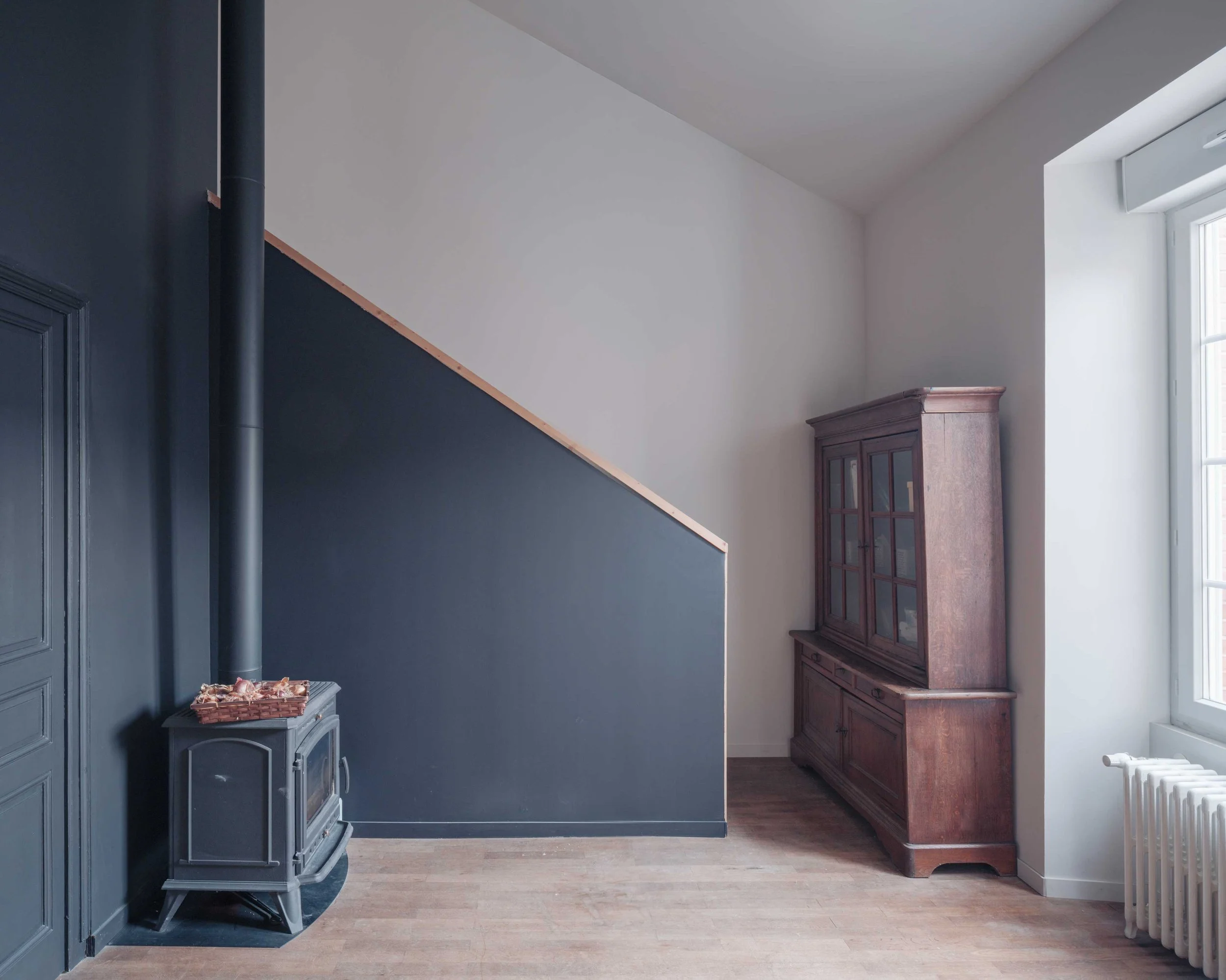 Intérieur d'une pièce avec un mur peint en bleu foncé sur la gauche, un mur blanc sous un plafond en pente sur la droite, une cheminée en bois ancienne et une grande fenêtre laissant entrer la lumière naturelle.