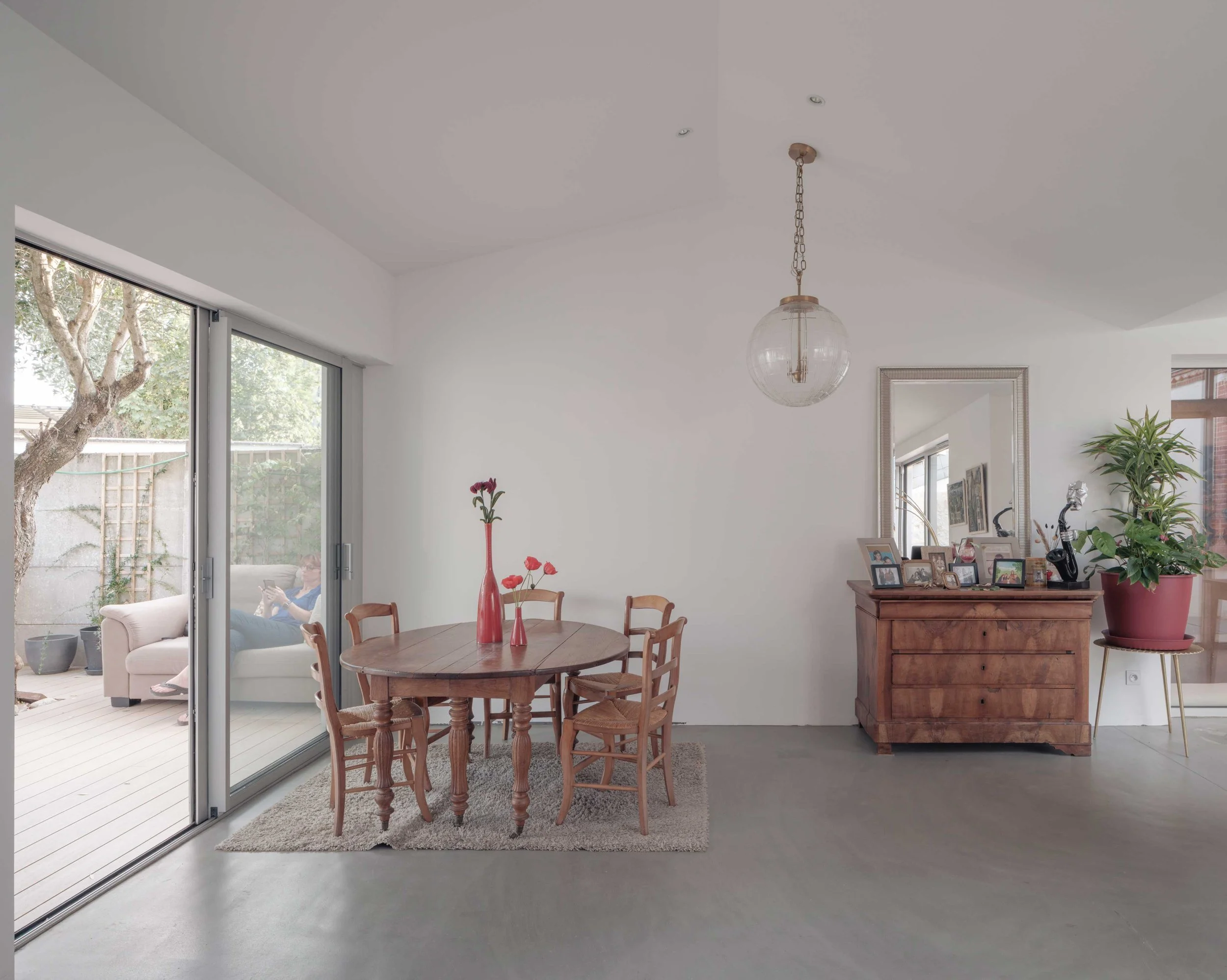 Salle à manger avec un meuble en bois, une table ronde en bois avec des chaises, une lampe suspendue en verre, des plantes, un miroir, et une large baie vitrée donnant sur une terrasse extérieure avec un arbre visible.