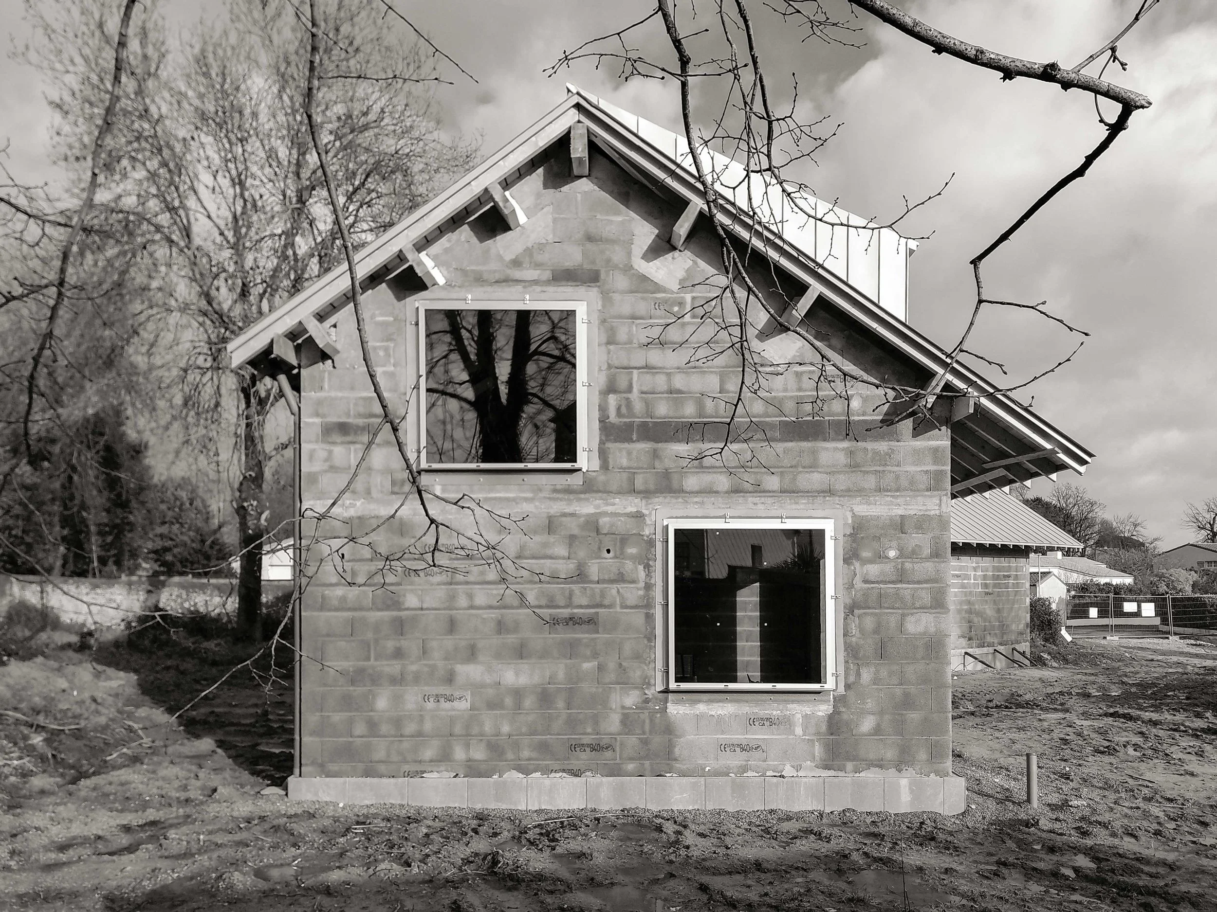 Une maison en construction en pierre, sans peinture, vue de face. La maison possède deux fenêtres sans volets et un toit en pente, avec quelques branches d'arbres dénudés visibles en premier plan.