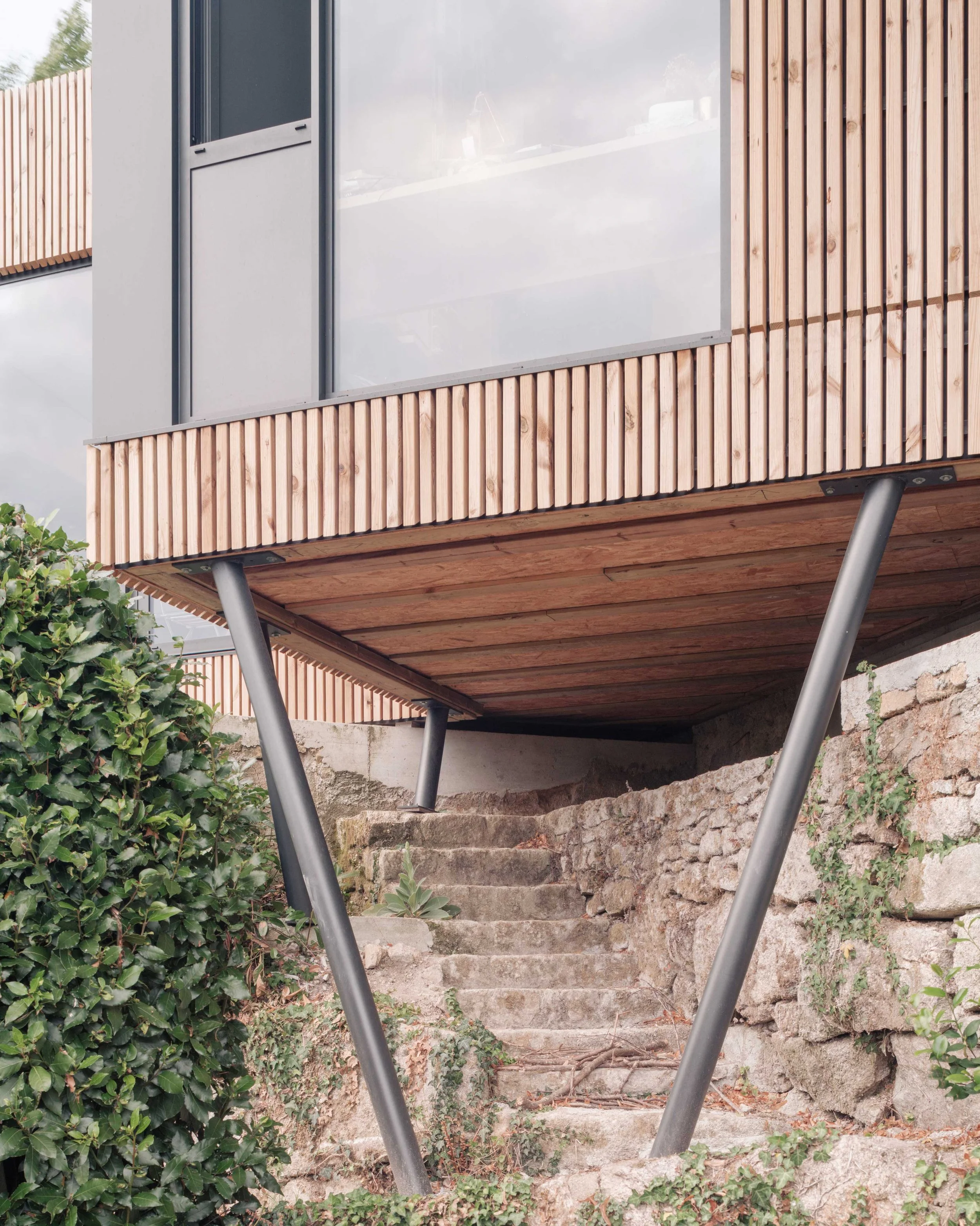 Escalier en pierre menant à une porte en verre avec des panneaux en bois, entouré de végétation et de murs en pierre.