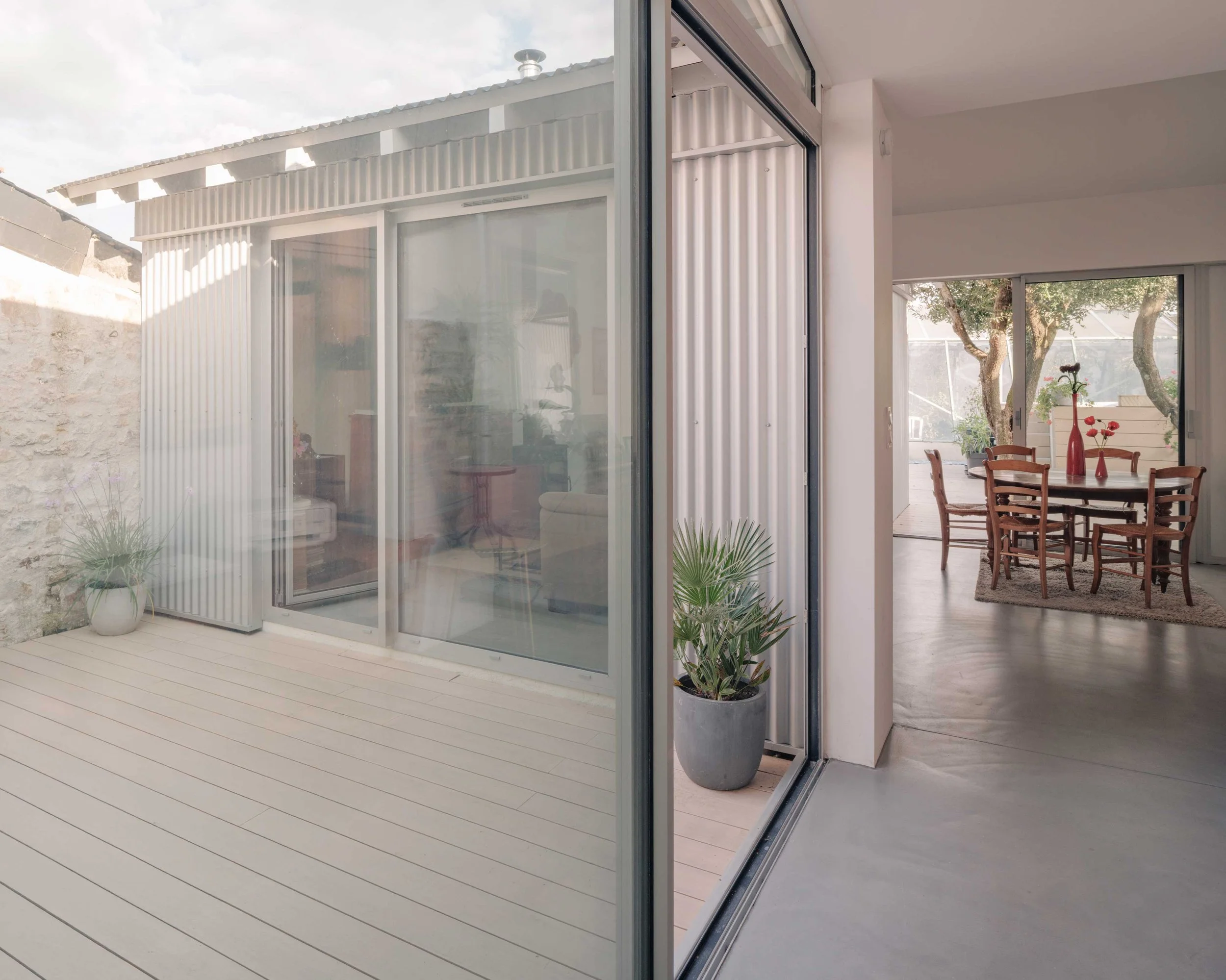 Intérieur d'une maison avec une pièce à vivre lumineuse comprenant une table en bois avec des chaises, des plantes en pot, et une grande baie vitrée laissant entrer la lumière naturelle, permettant de voir un extérieur avec des arbres.
