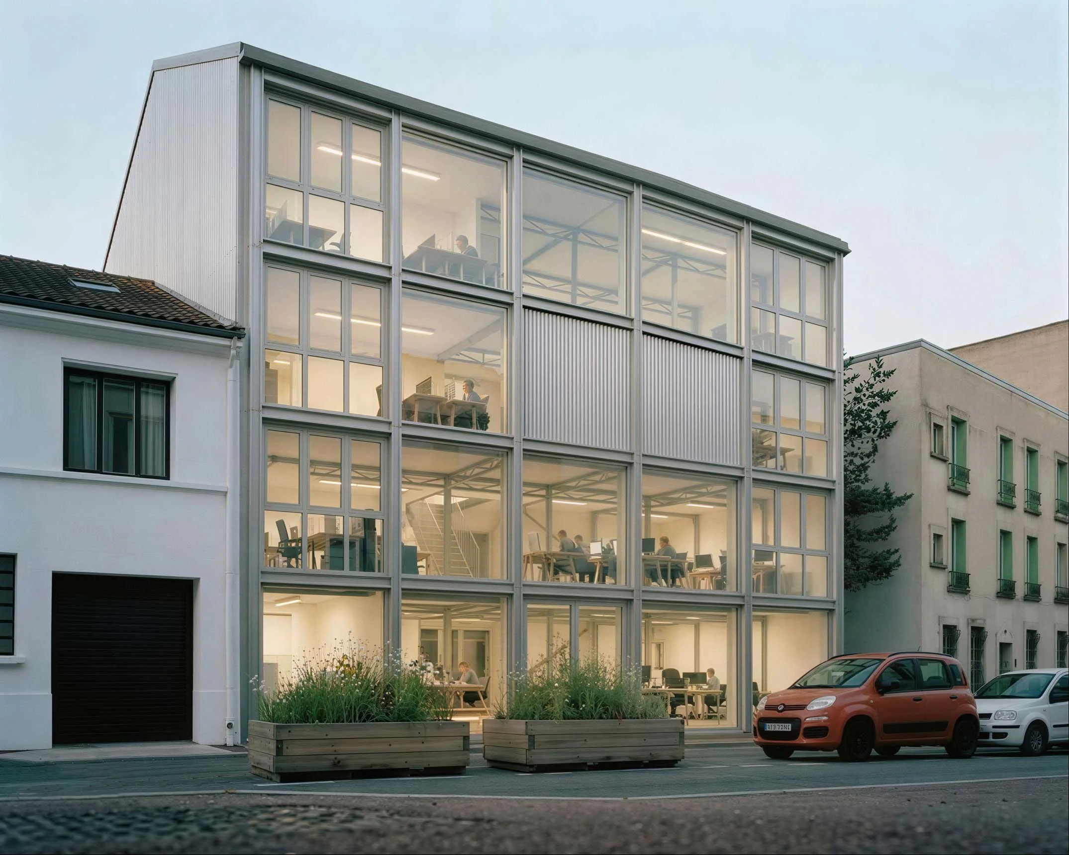 Bâtiment moderne avec façade en verre, où l'on voit des personnes travaillant à l'intérieur. Il y a des voitures garées devant et des jardinières en bois avec des plantes, sous un ciel clair.