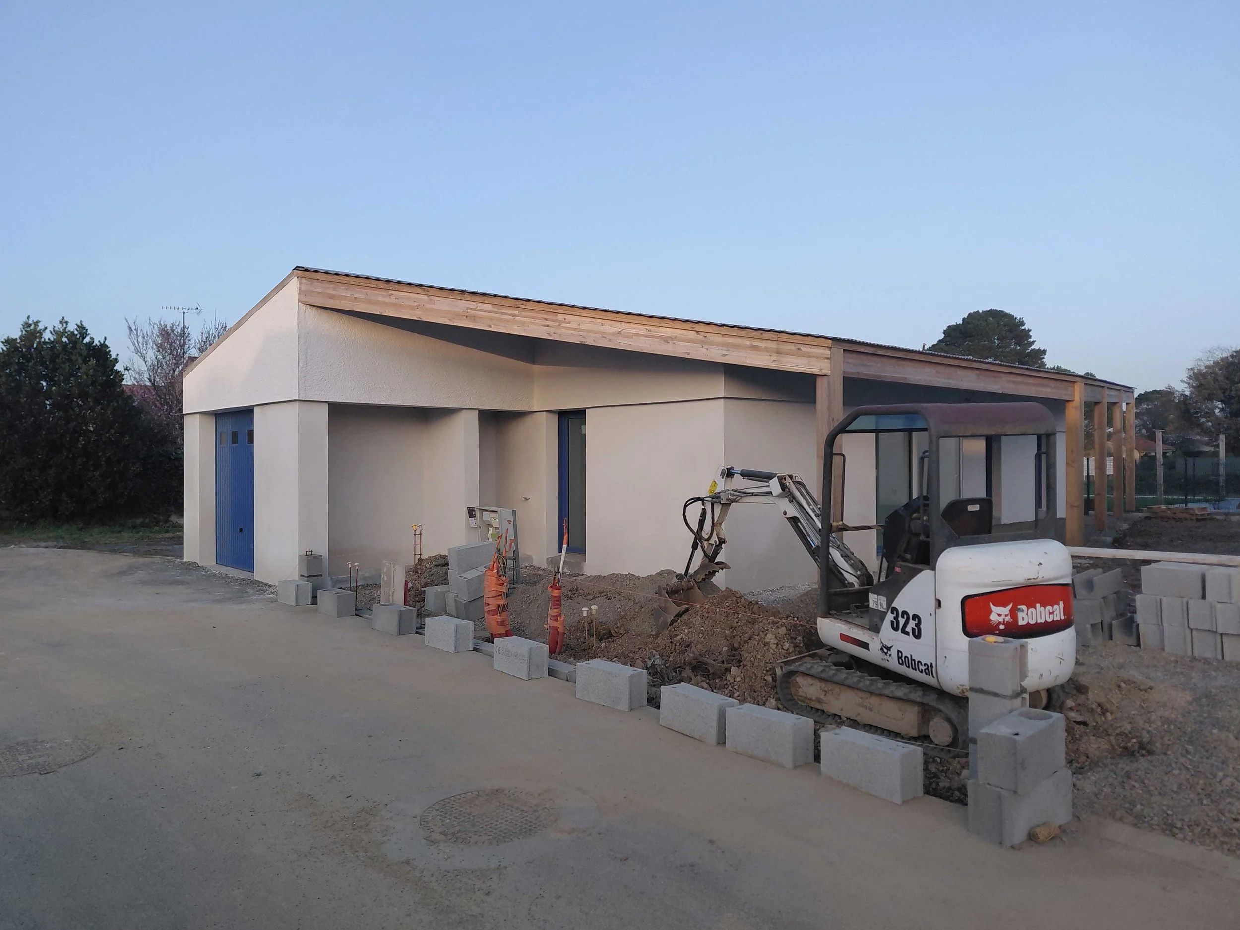 Construction de maison moderne avec mur blanc, toiture en bois, et grande baie vitrée, chantier en cours avec machine Bobcat et blocs de béton, en extérieur avec arbres et ciel clair.