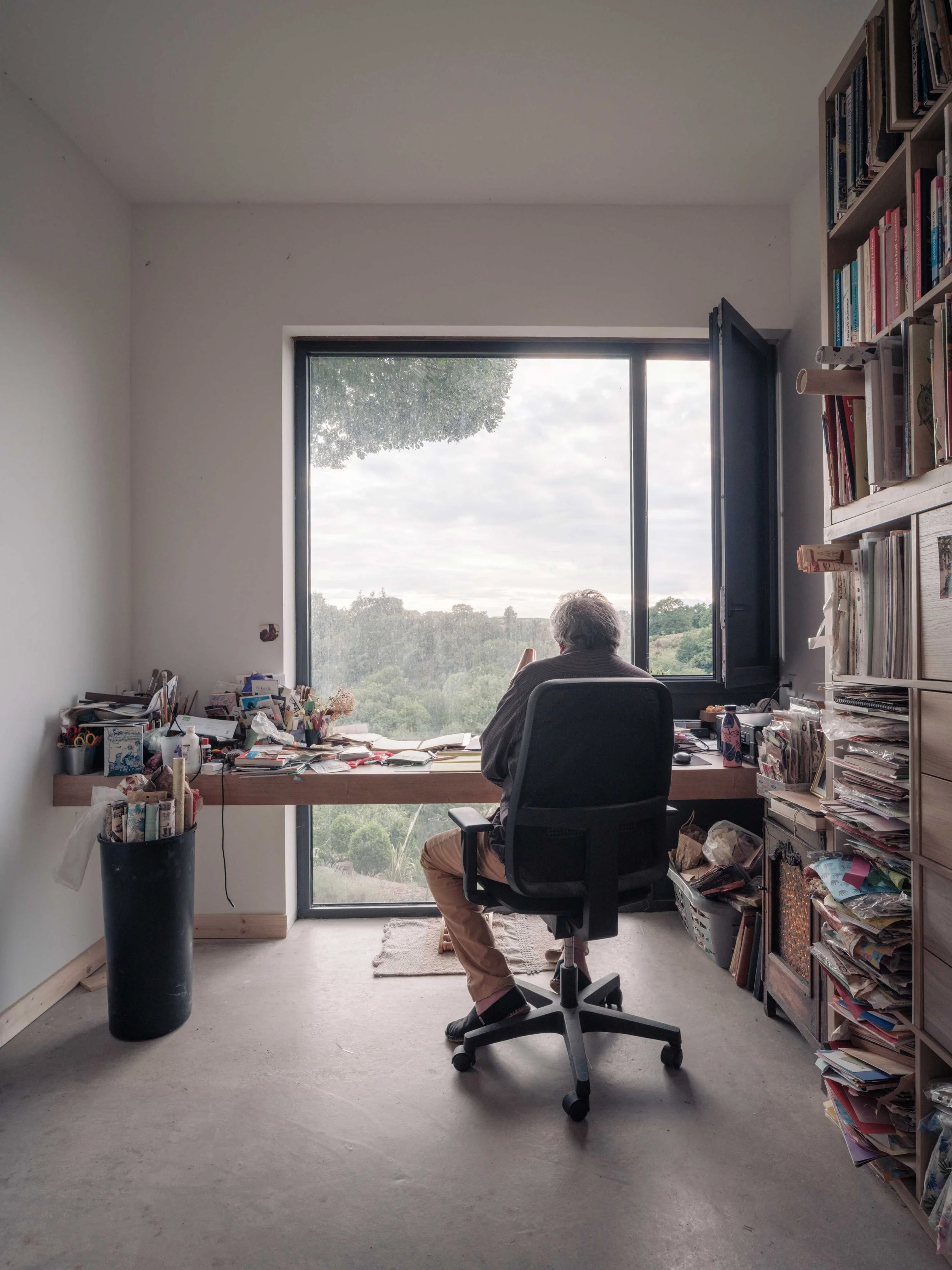 Un bureau avec un homme âgé assis devant une grande fenêtre ouverte, entouré de livres, papiers, et fournitures de bureau.