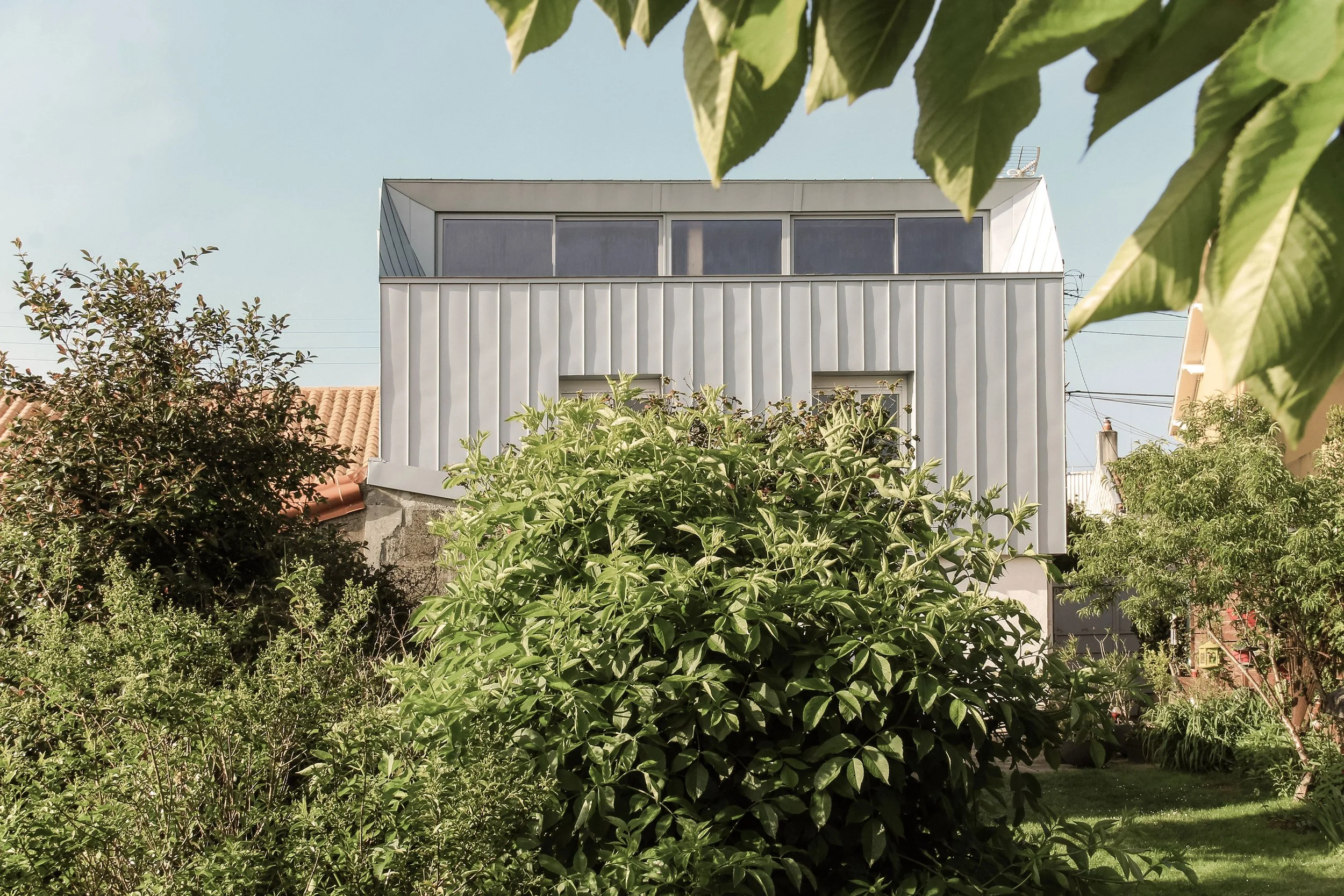 Une maison moderne à plusieurs étages, entourée de végétation luxuriante avec des arbres et des buissons, sous un ciel clair.