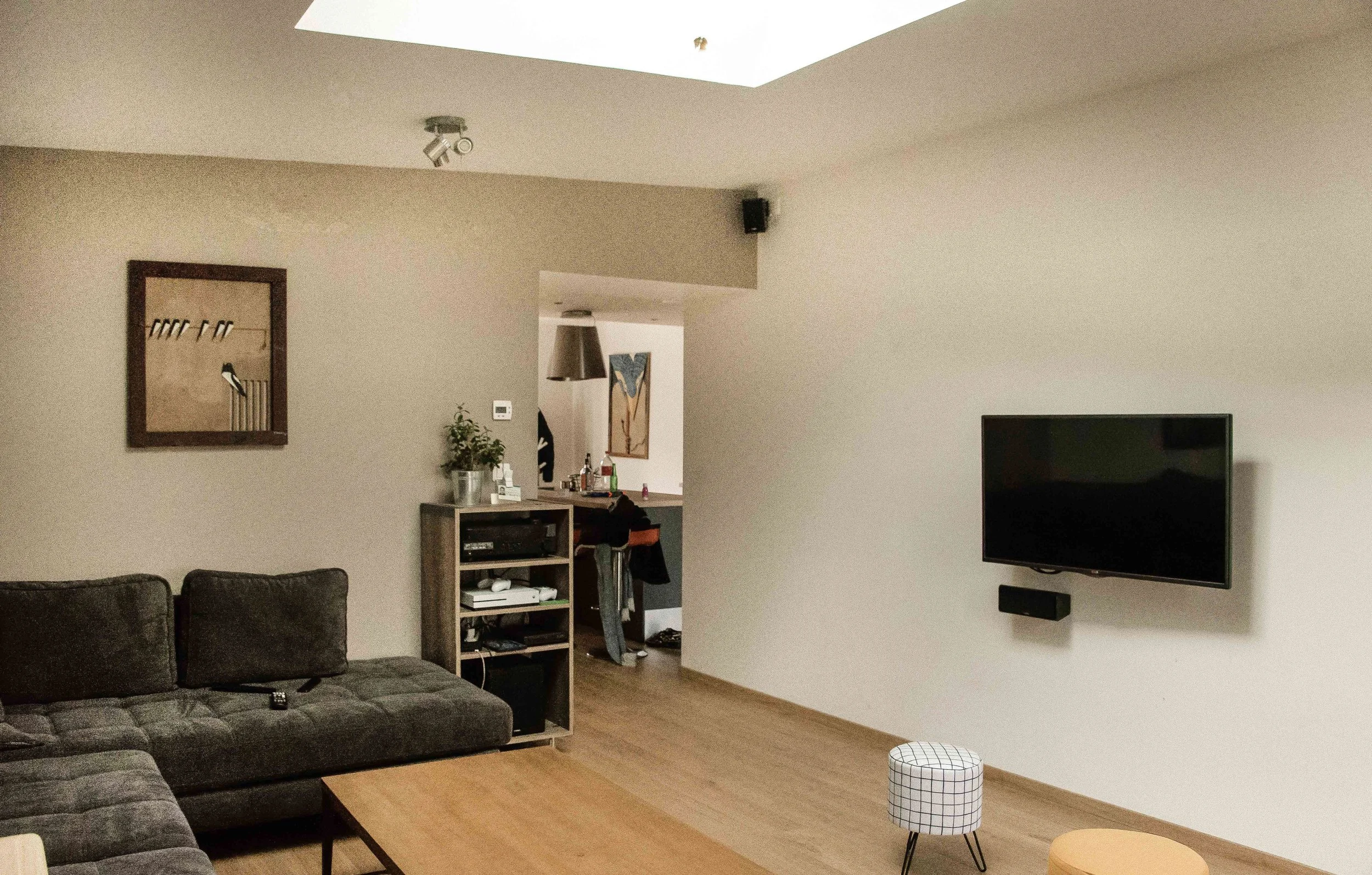 Salon moderne avec fauteuils en tissu gris, télévision à écran plat sur le mur, sculpture abstraite dans la pièce adjacente, et plantes décoratives. La pièce a un plafond avec une ouverture lumineuse naturelle.