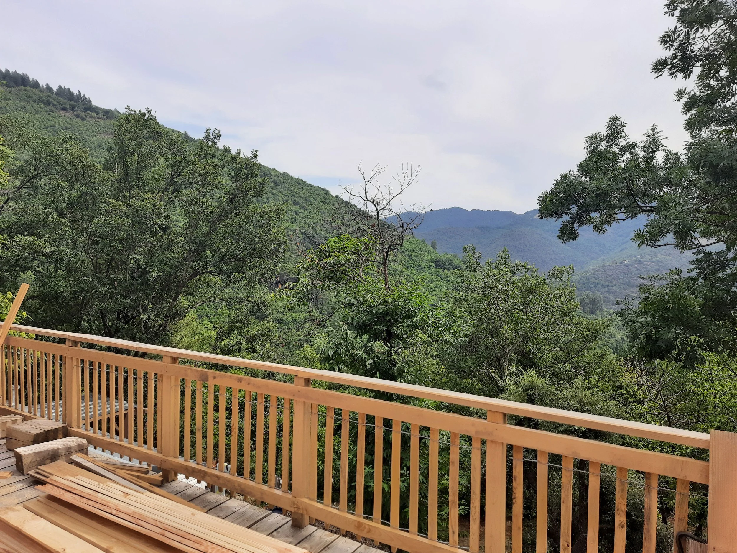 Vue d'une terrasse en bois en construction avec un garde-corps en bois et un paysage montagneux et forestier en arrière-plan.