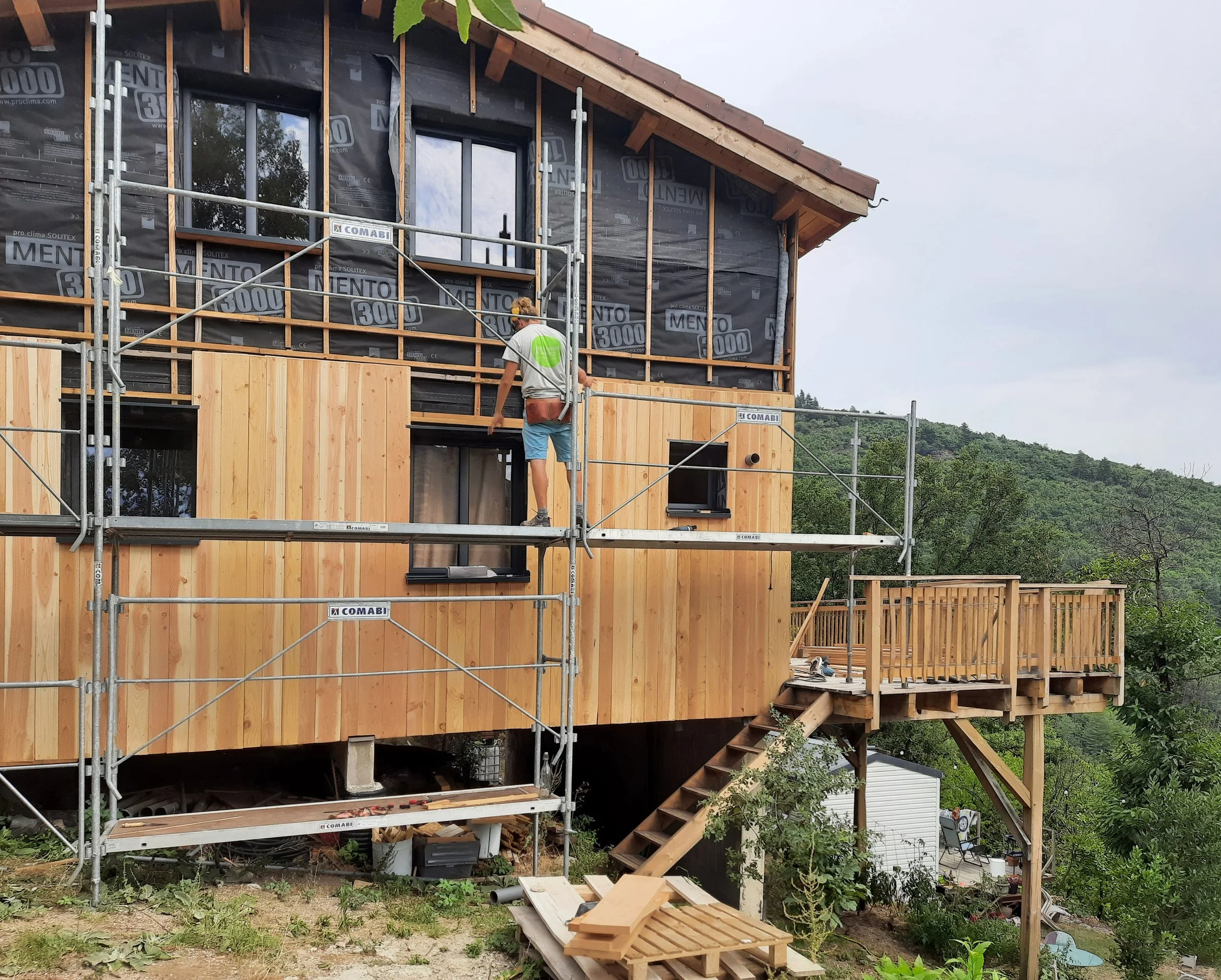 Une maison en construction avec des murs enbois et des fenêtres, entourée d'un échafaudage, dans un paysage verdoyant avec des collines en arrière-plan.