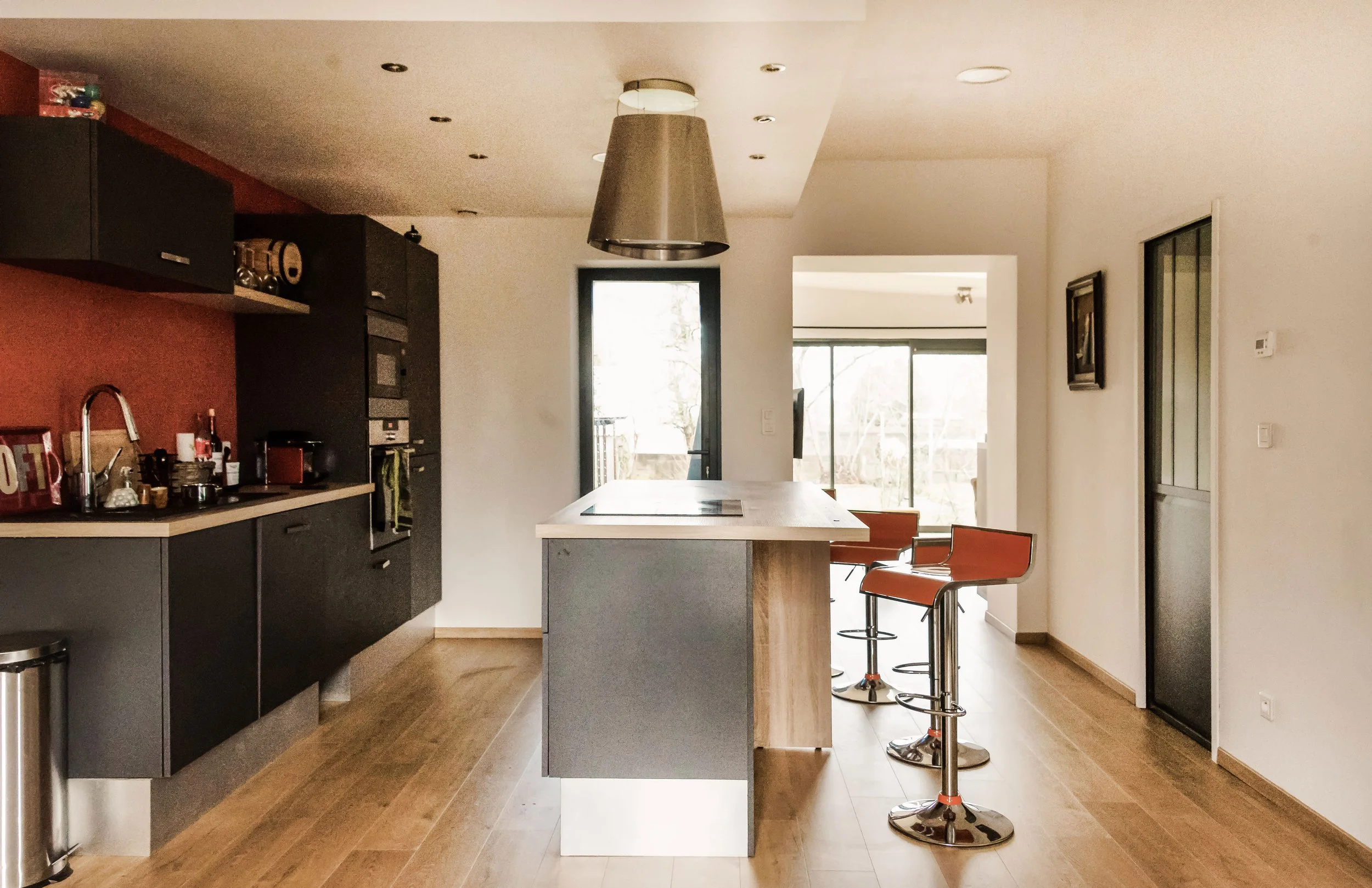 Cuisine moderne avec un îlot central, chaises hautes orange, fenêtres donnant sur un balcon ou un jardin, et éclairage encastré au plafond.