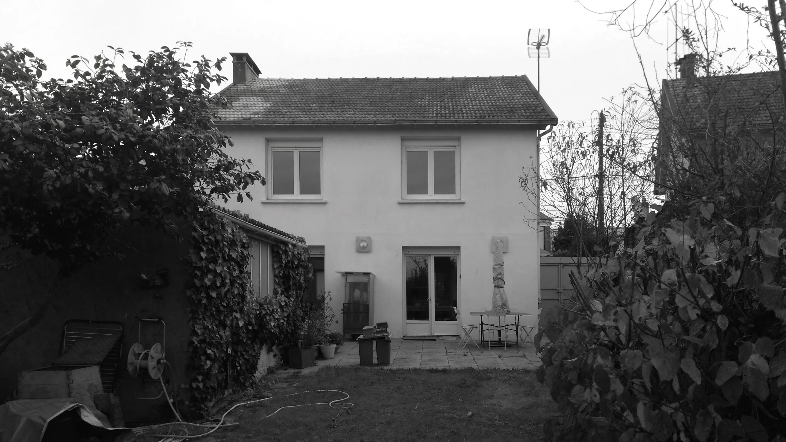 Une maison à deux étages avec un toit en tuiles, vue du jardin arrière, avec des fenêtres, une porte-fenêtre, des meubles de jardin, et des arbres dénudés, le tout en noir et blanc.